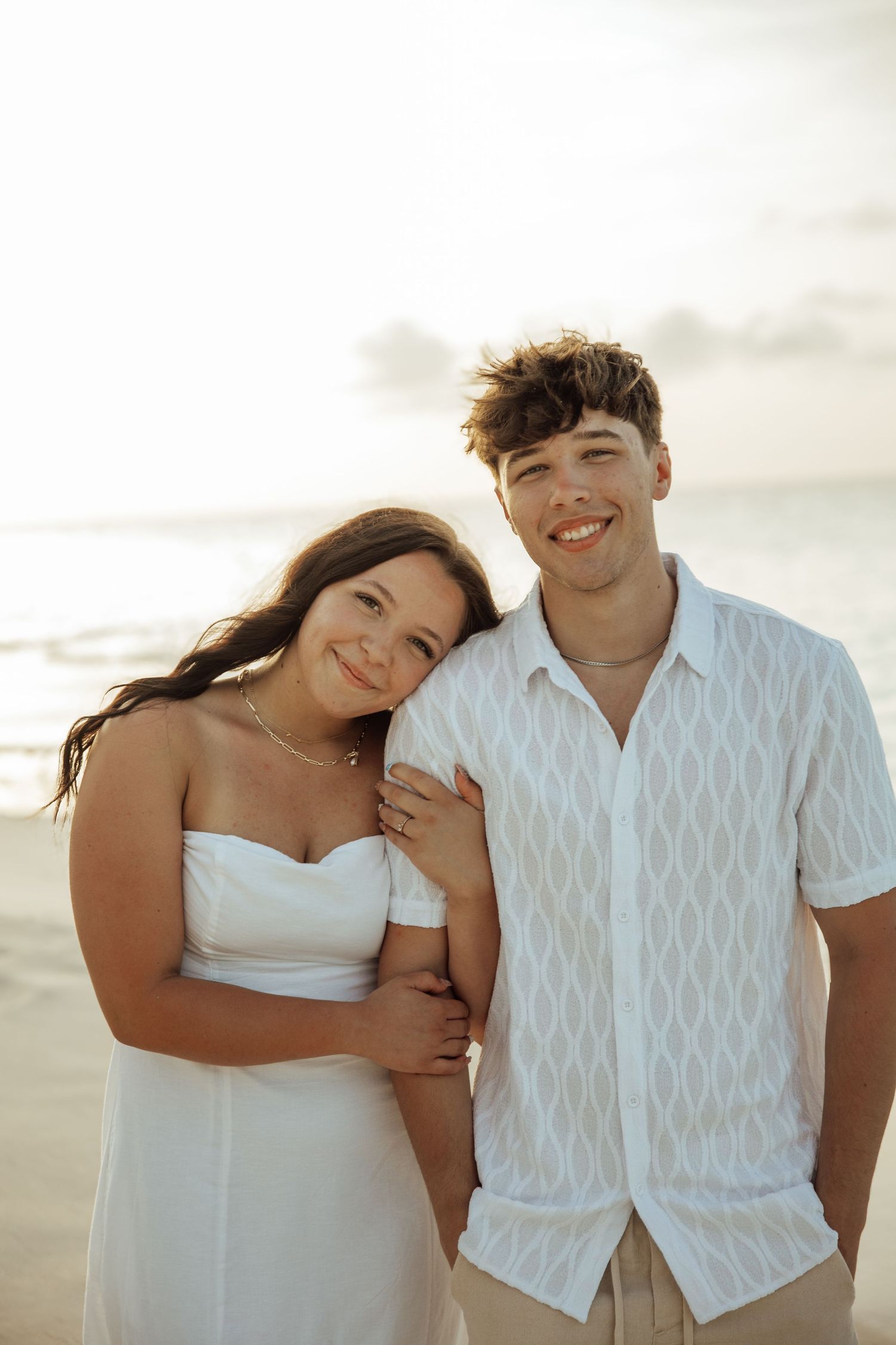 Smiling couple posing together on a beach at sunset, both dressed in white outfits with ocean backdrop.