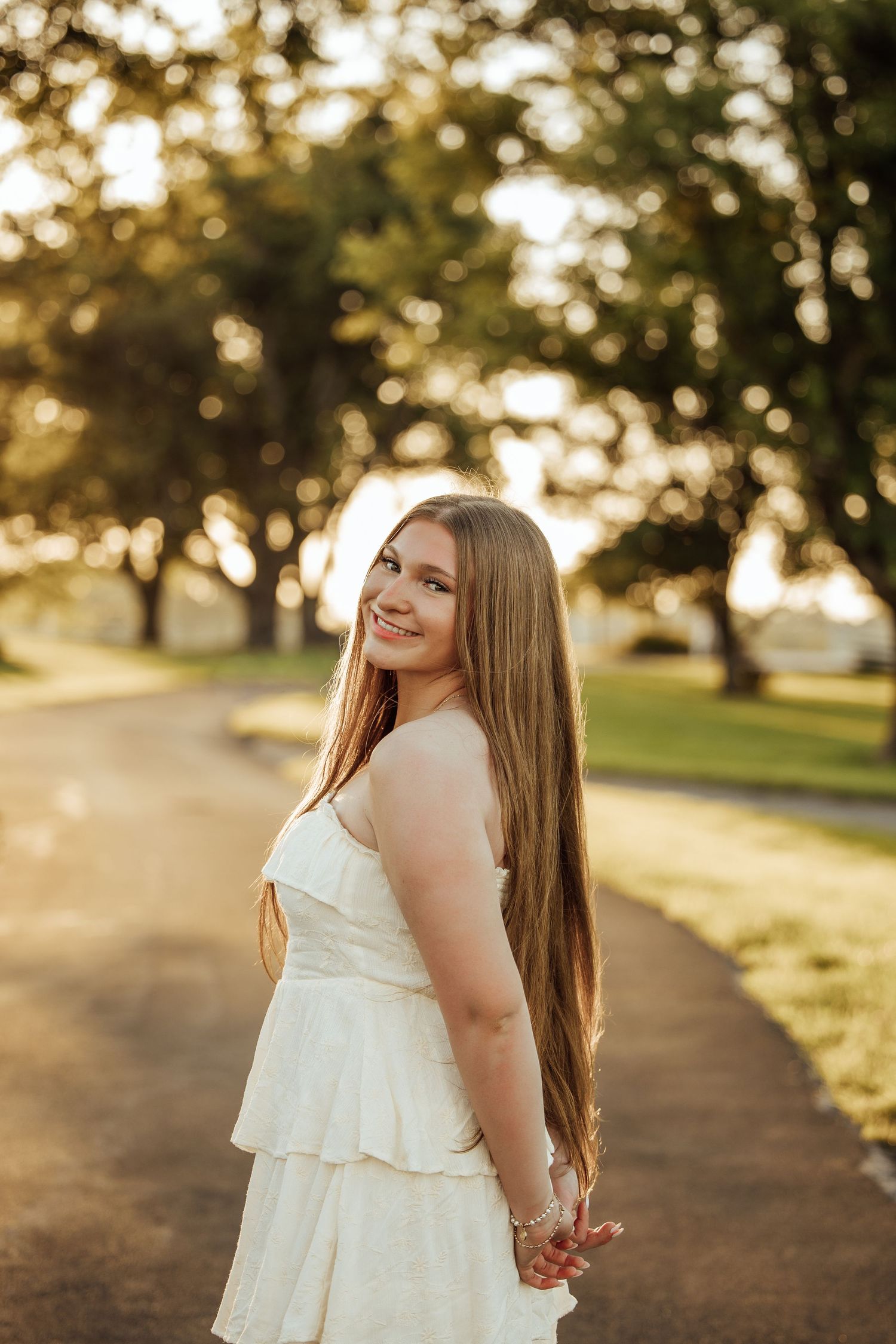 Young woman in white dress smiling over shoulder on sunlit park path with golden bokeh trees.