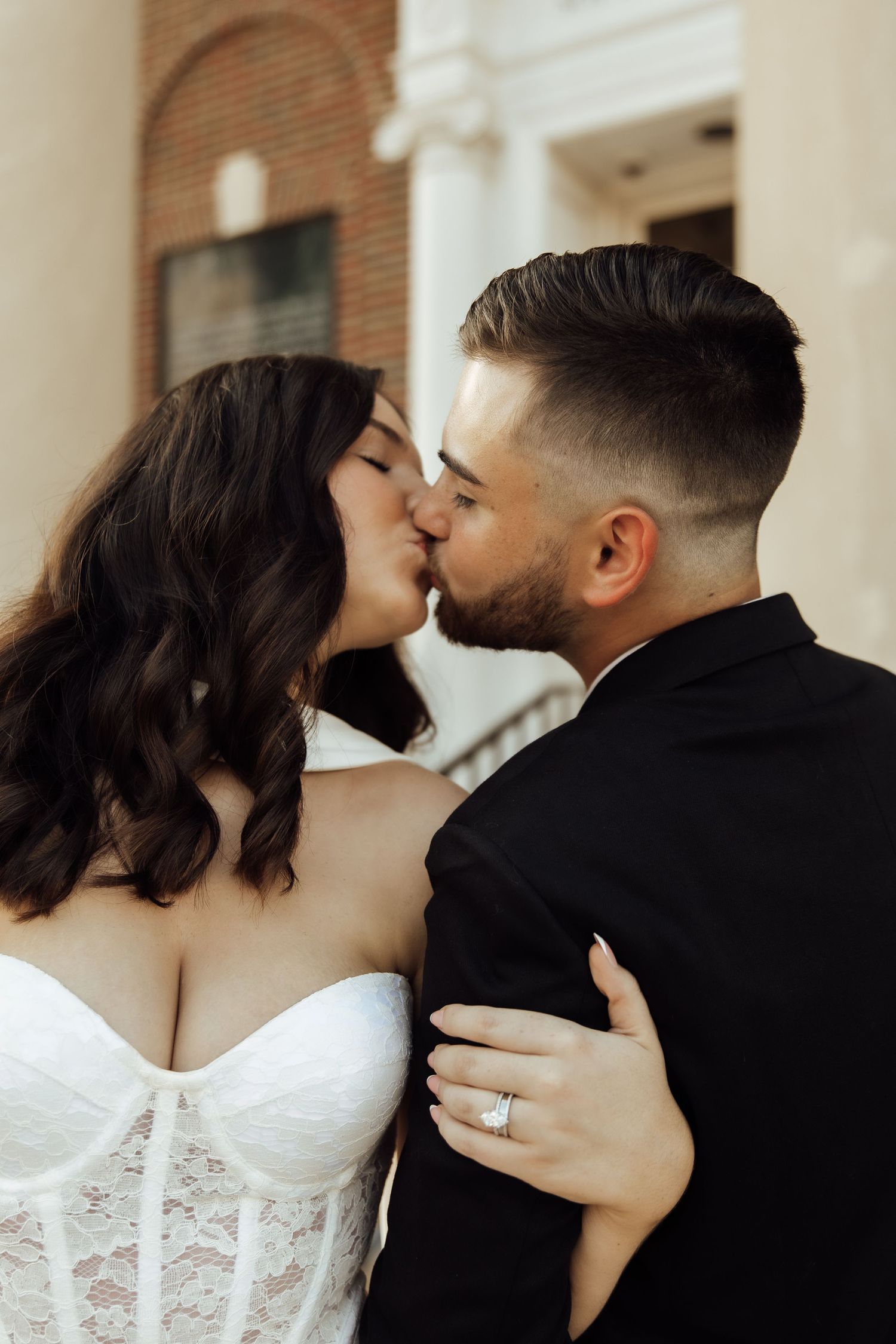 A bride and groom share a romantic kiss, she wears a white lace corset gown and diamond ring, he wears a black suit.