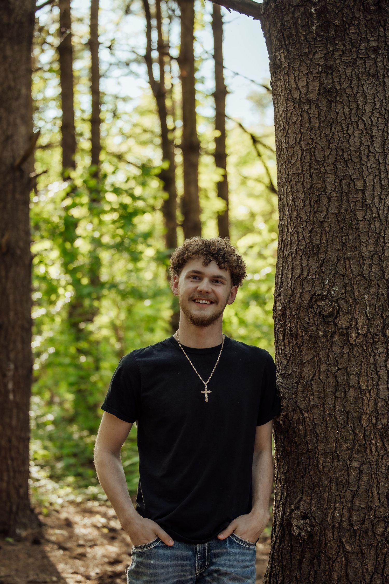 Smiling young man in black t-shirt with gold cross necklace stands between trees in forest.