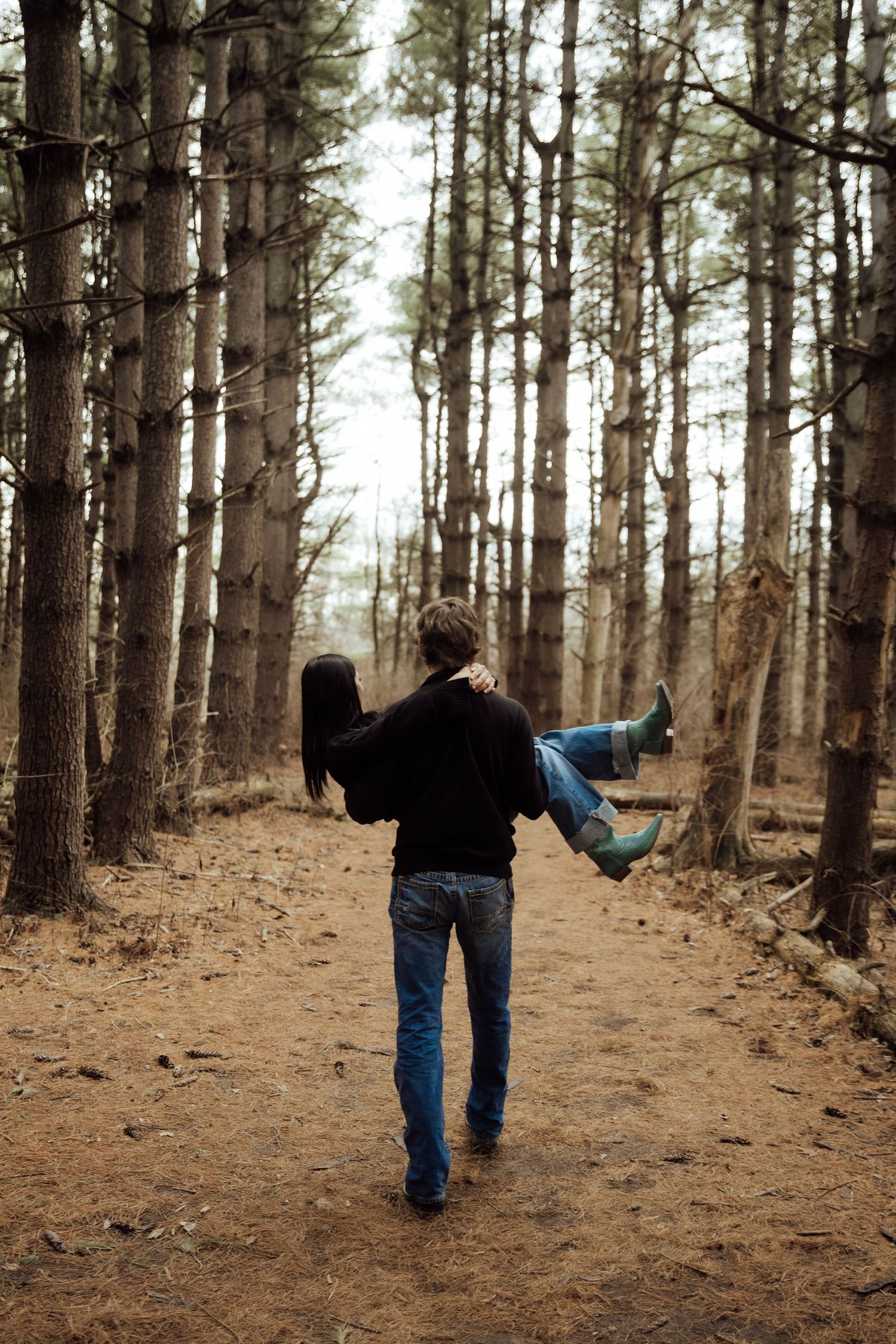 Man carrying woman in his arms while walking down a scenic forest path lined with tall trees.