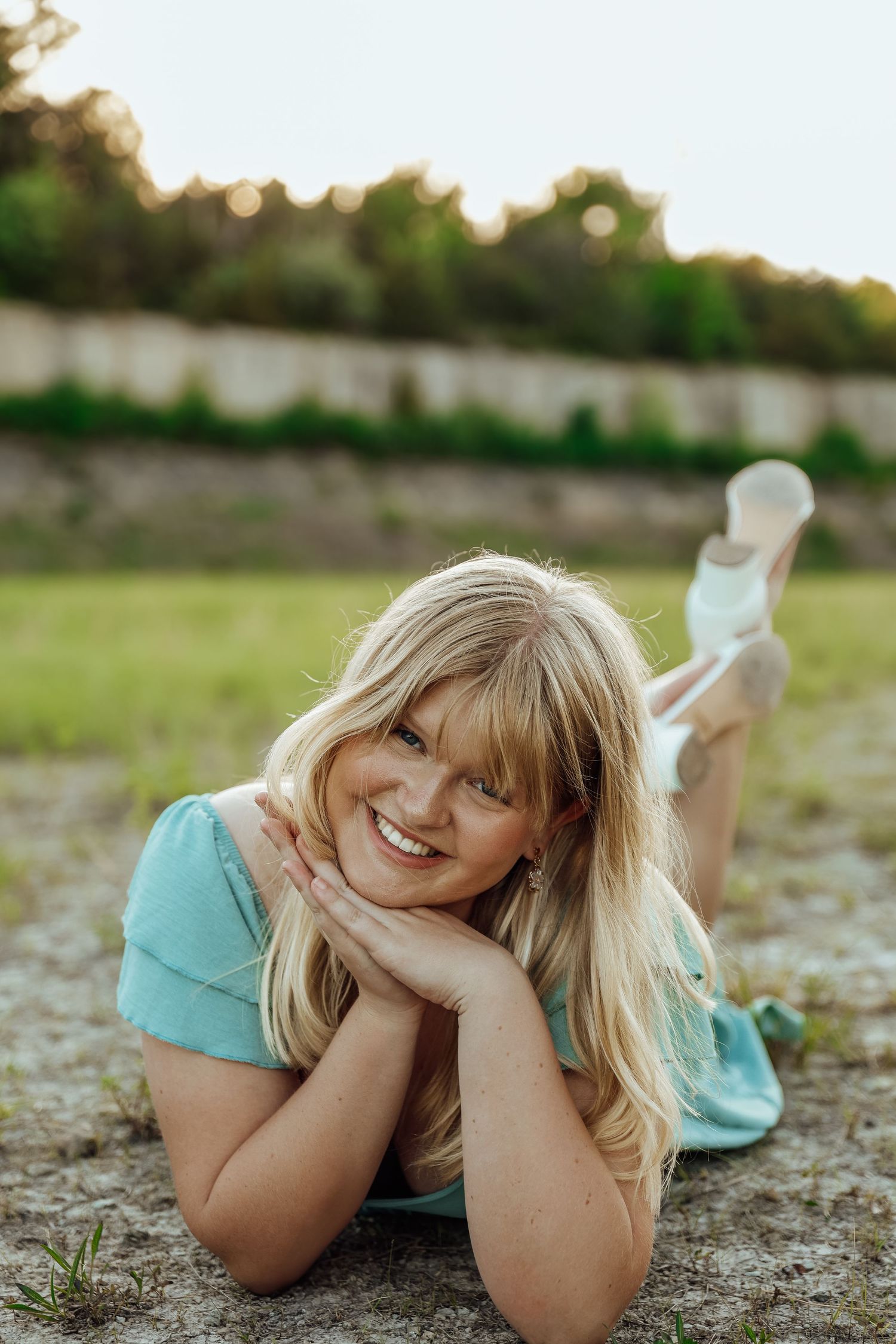 Smiling blonde woman in mint dress lying on ground outdoors in warm sunlight.