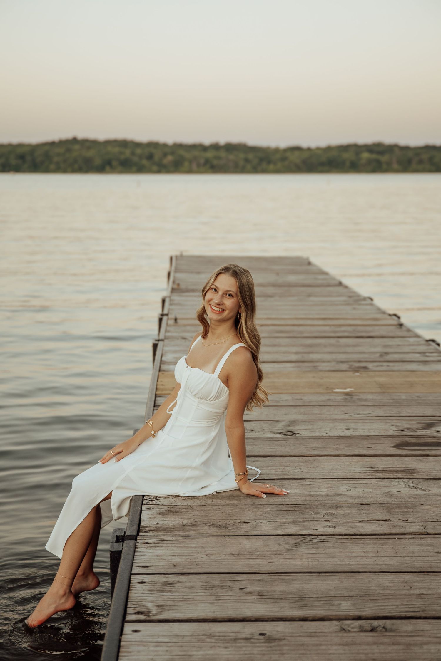 Smiling blonde woman in white dress sitting on wooden dock with legs dangling over calm lake.