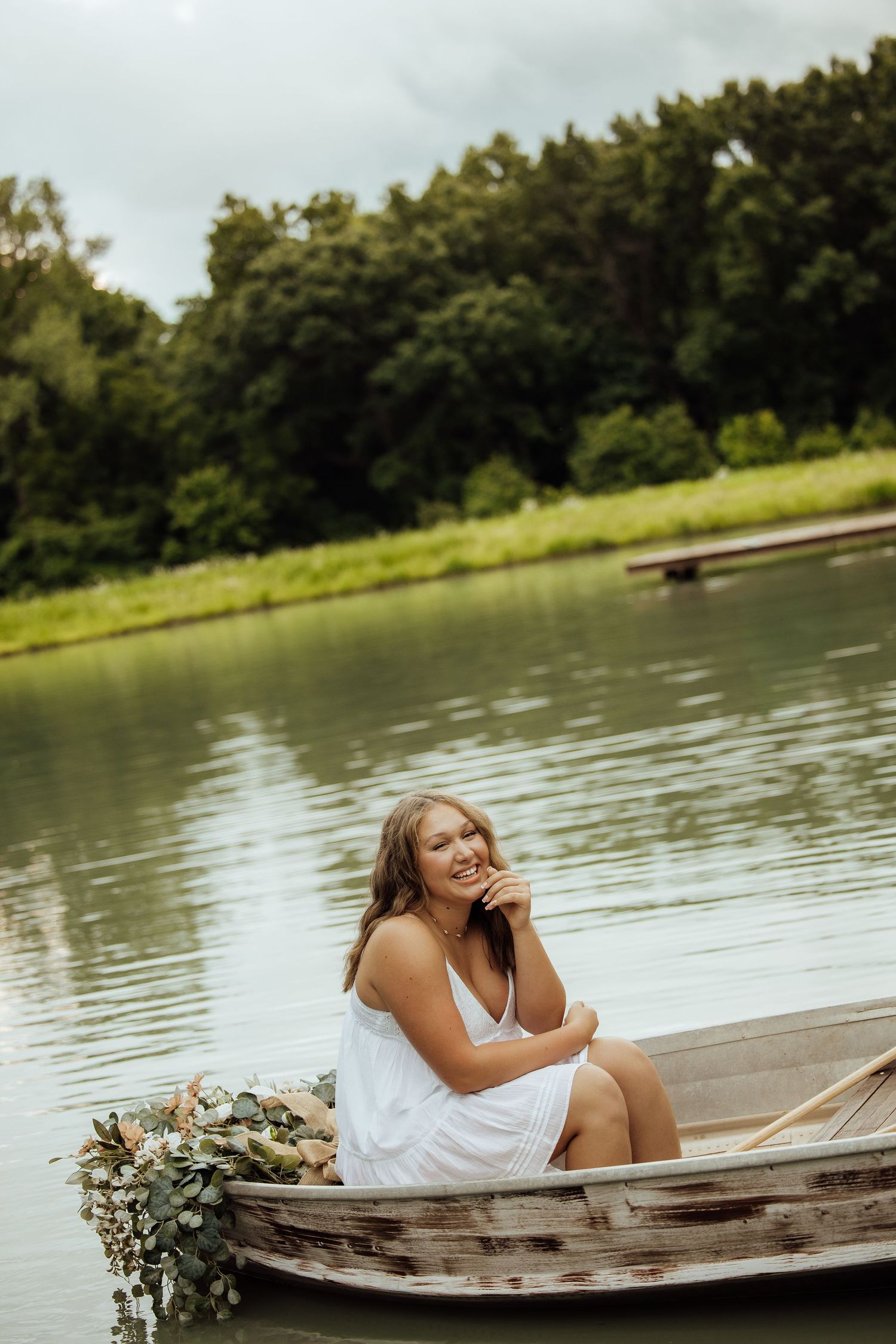 Young woman in white dress sitting in a rustic wooden boat with flowers on a calm lake surrounded by lush greenery.