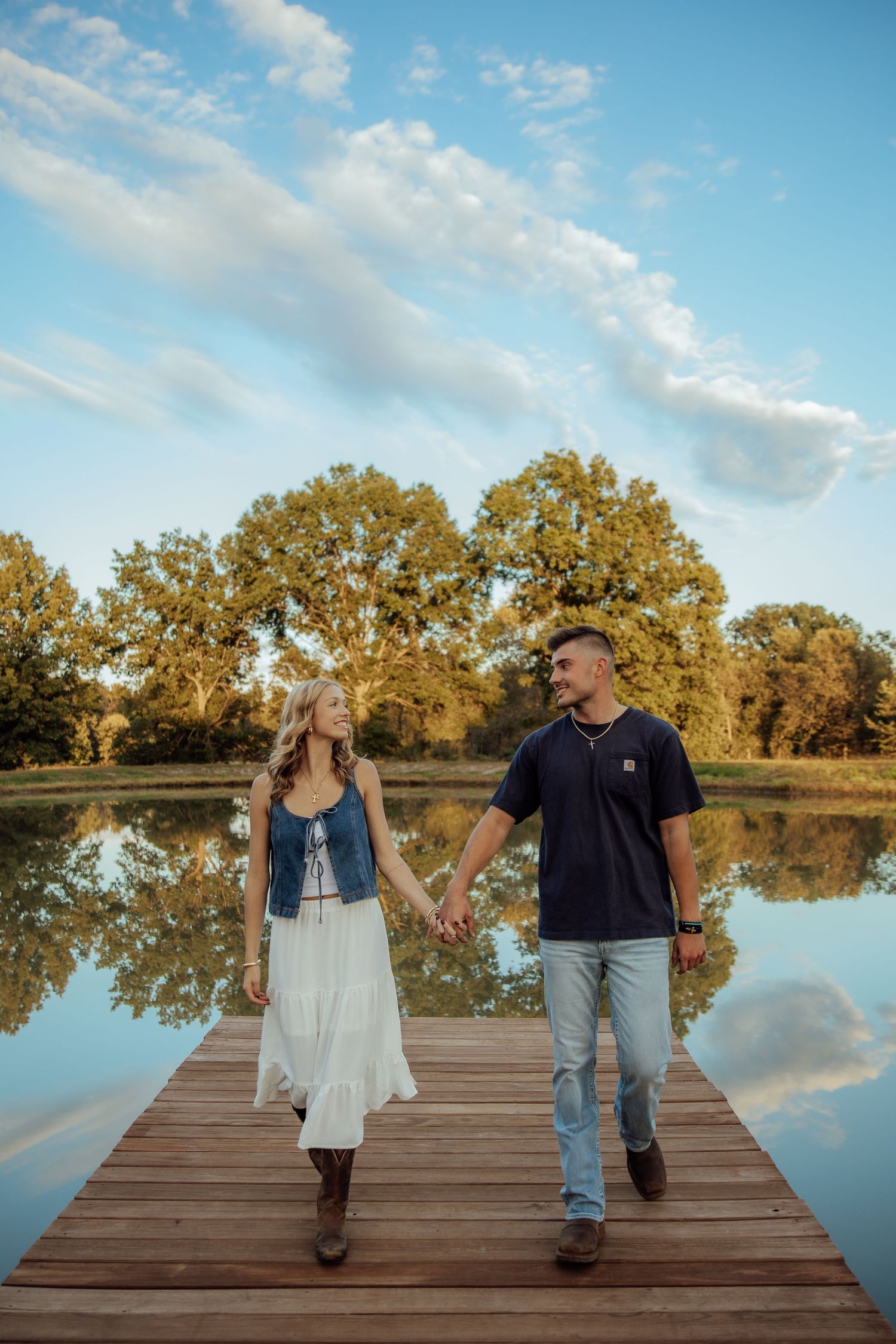 Couple holding hands on wooden dock by reflective lake, woman in white dress and denim vest, man in navy shirt.