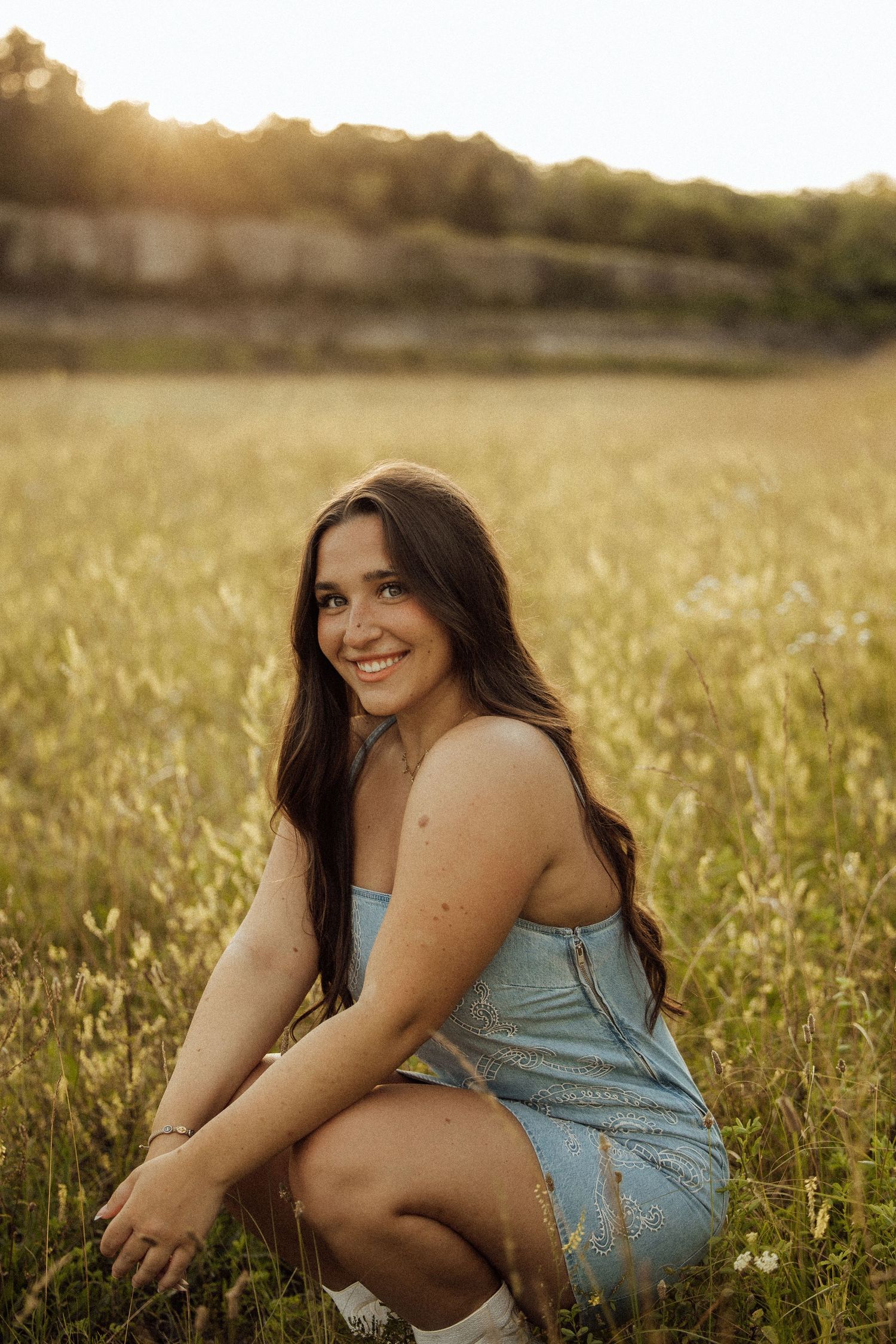 Young woman in blue denim dress crouching in golden field at sunset, smiling warmly at camera.