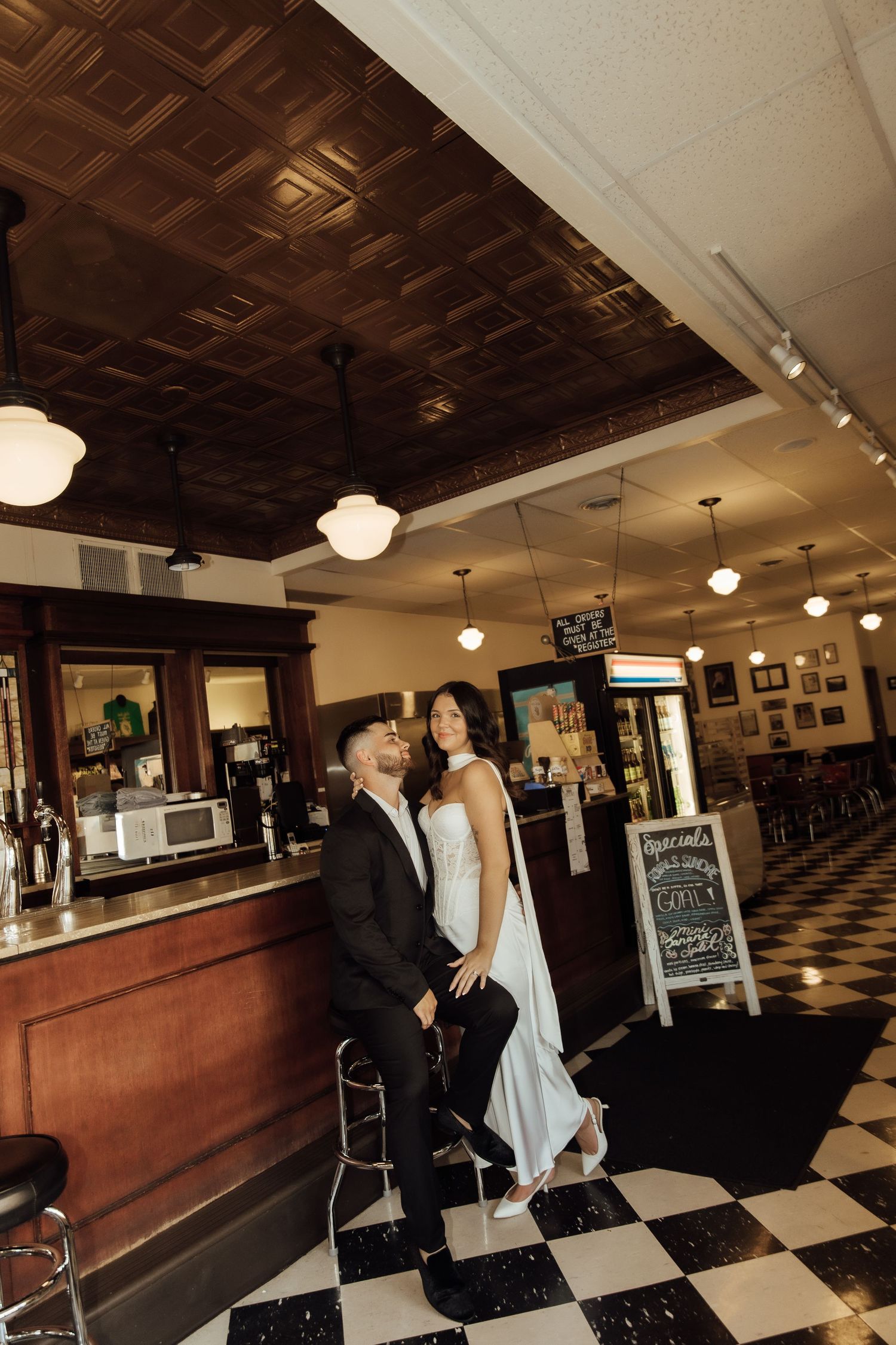 Elegant couple in formal attire posing at a vintage bar with checkered floors and ornate tin ceiling.