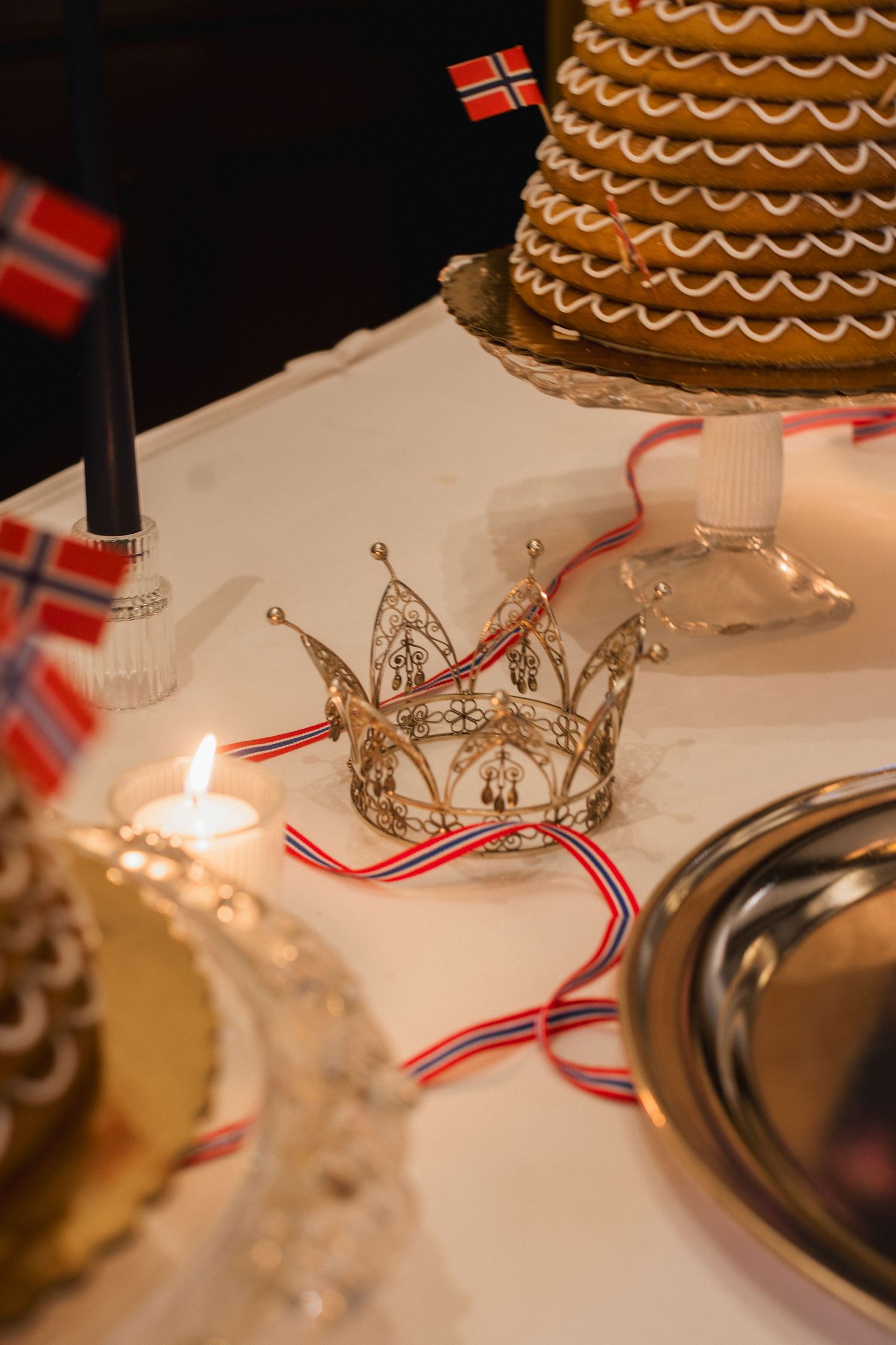Norwegian bridal crown with red and white ribbon displayed on a table beside a traditional kransekake wedding cake.