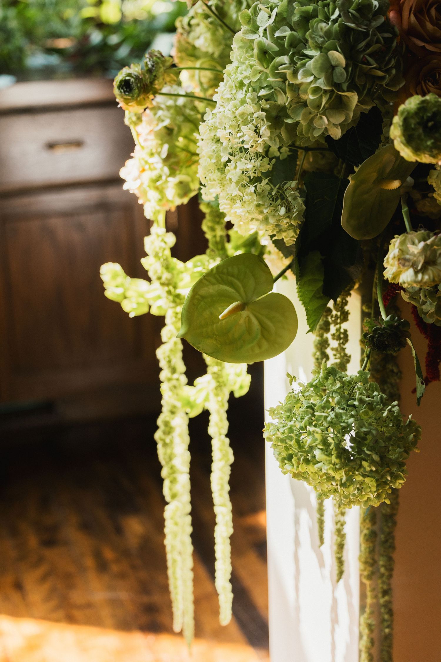 Lush green floral arrangement with hydrangeas, anthuriums, and hanging amaranthus decorating a white pillar indoors.
