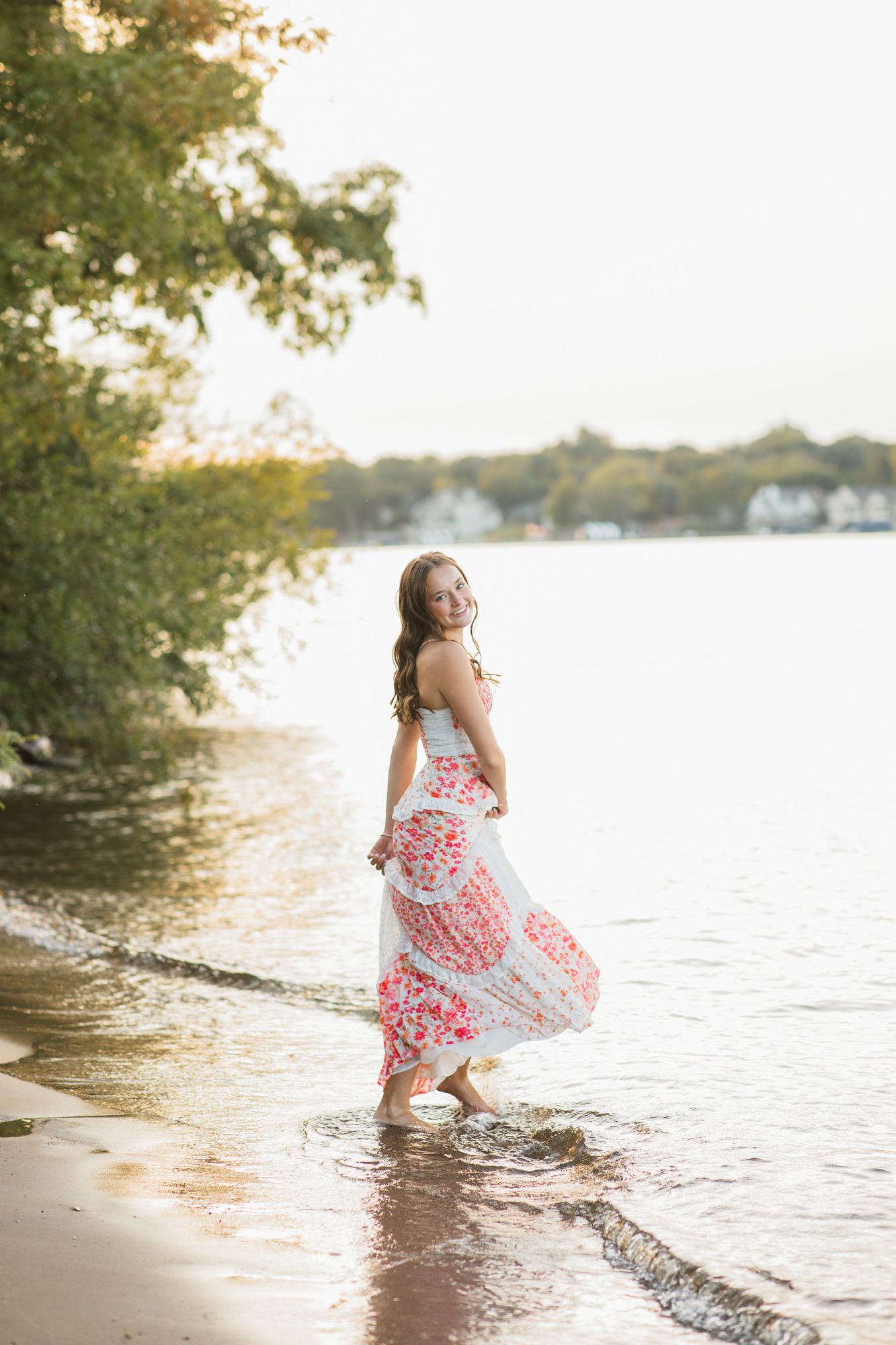Young woman in floral maxi dress wading barefoot in shallow lake water, smiling over shoulder at golden hour.