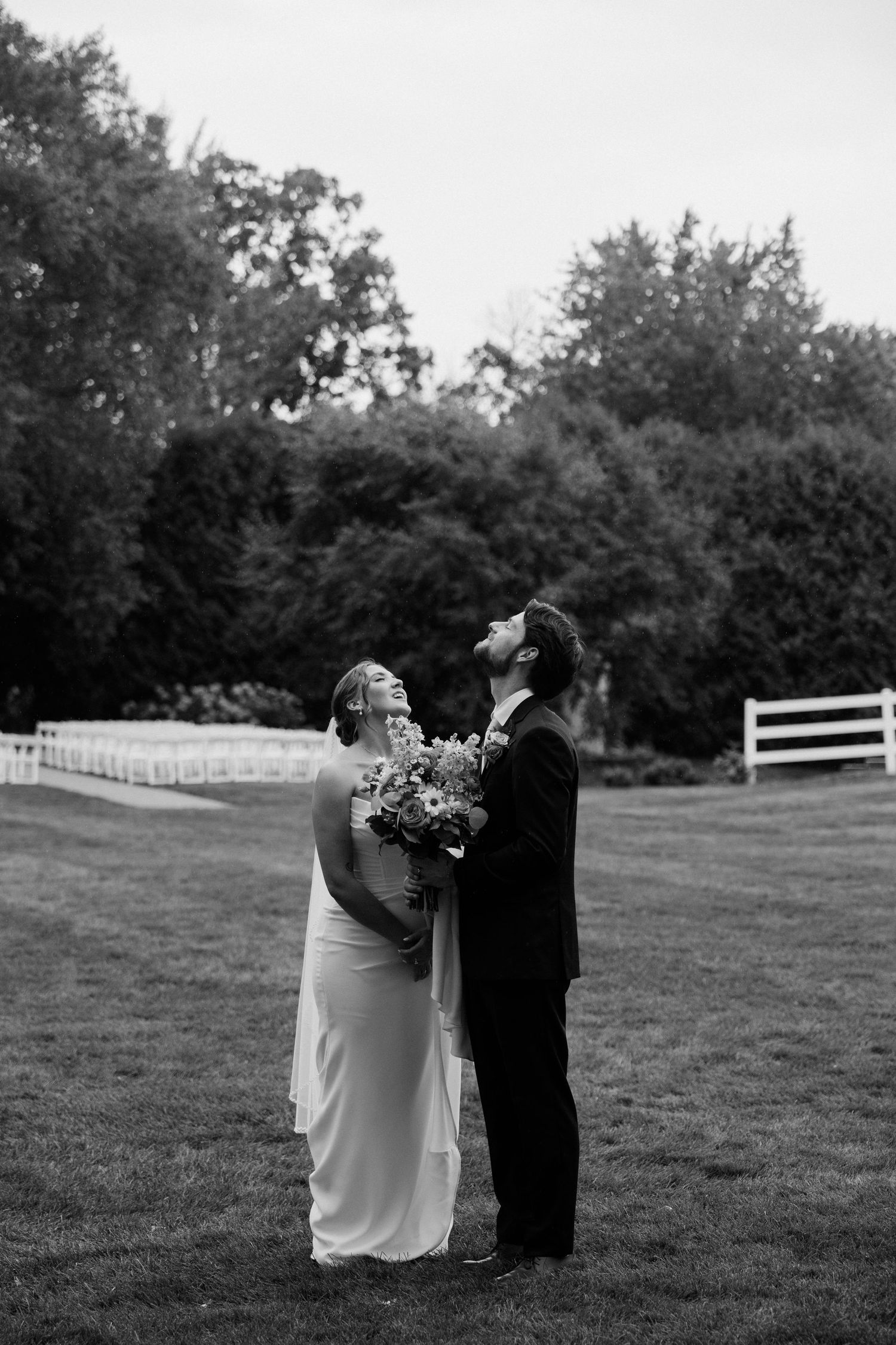 Black and white photo of a bride and groom standing on a lawn, both looking up, surrounded by lush trees and a white fence.