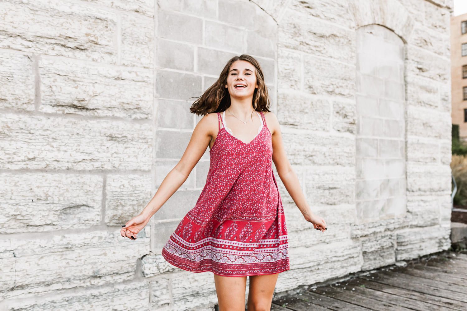 Person dancing joyfully in red summer dress against white stone wall.
