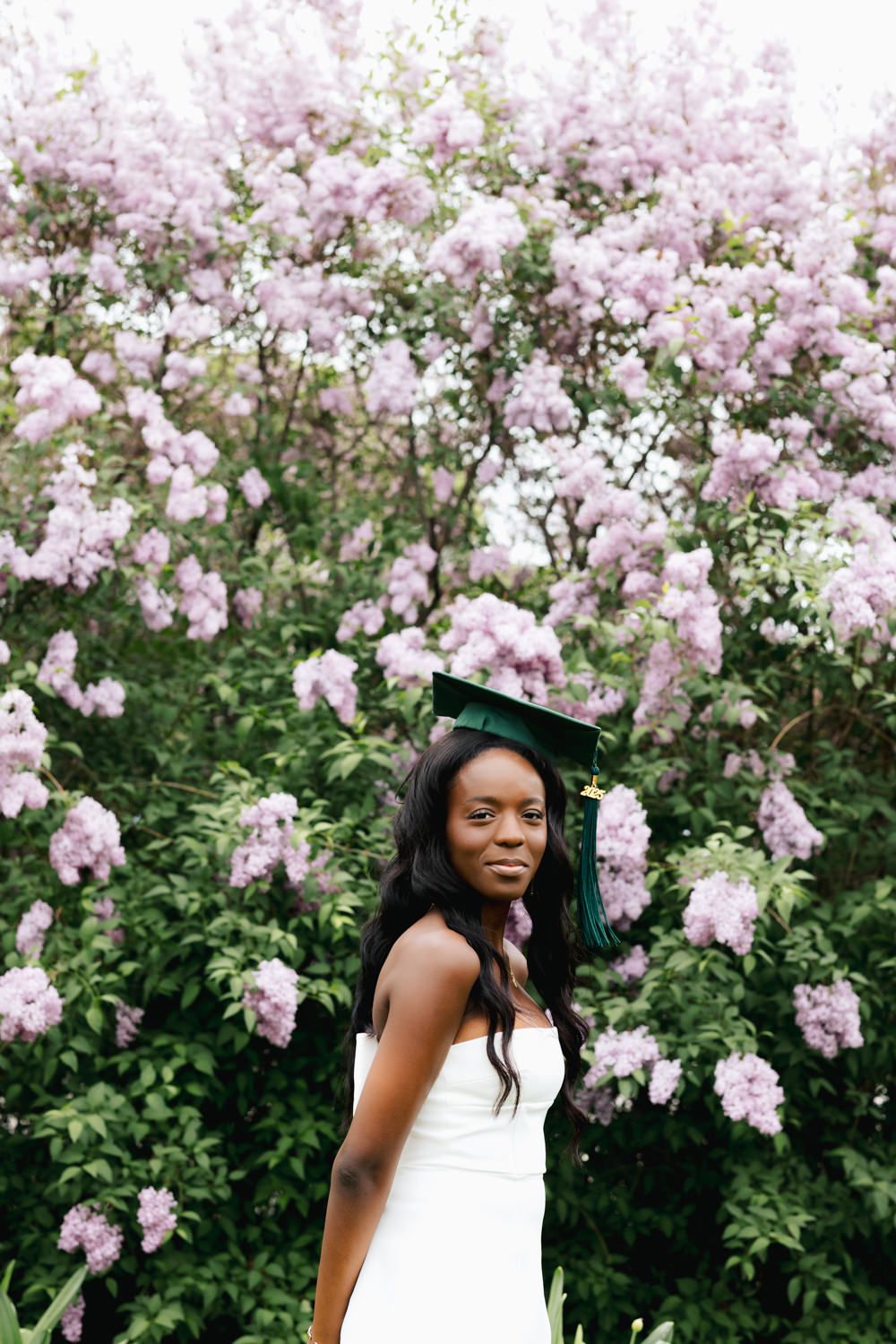 Graduate in white dress and green cap smiles among pink flowering trees.