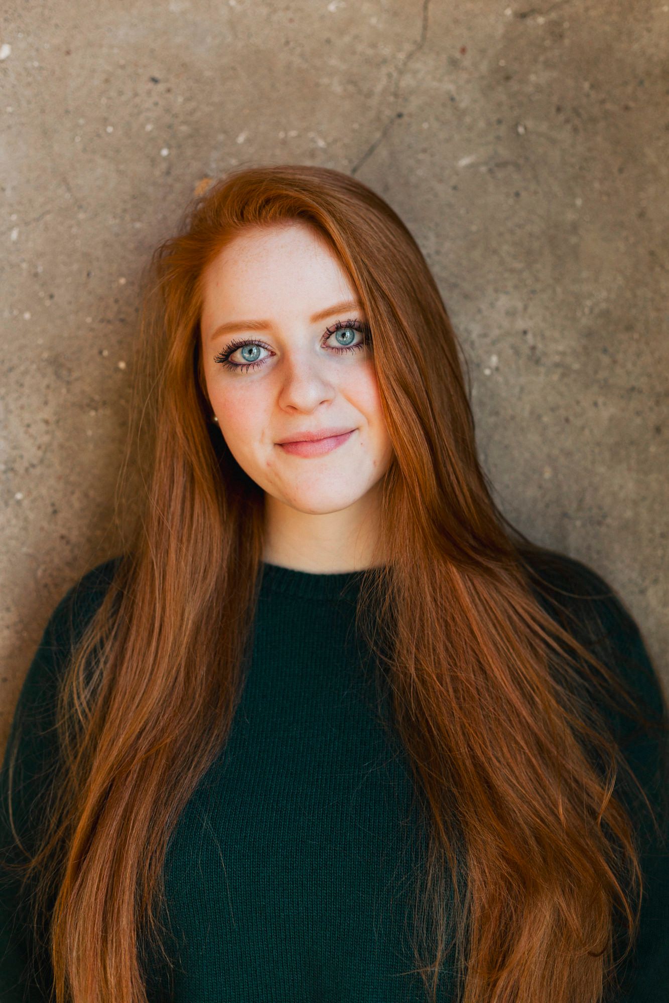 Portrait of a redhead in a dark green sweater against a concrete wall background.