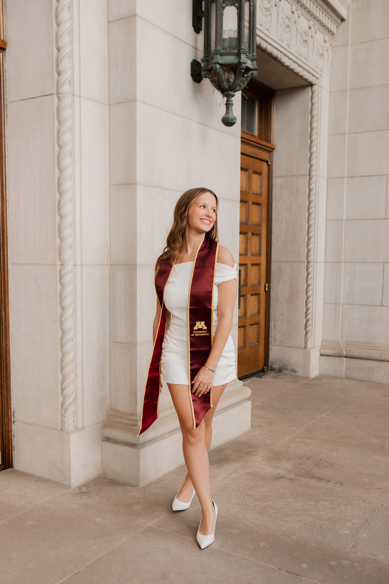 Young woman in white dress with maroon University of Minnesota graduation sash walking near wooden doors.