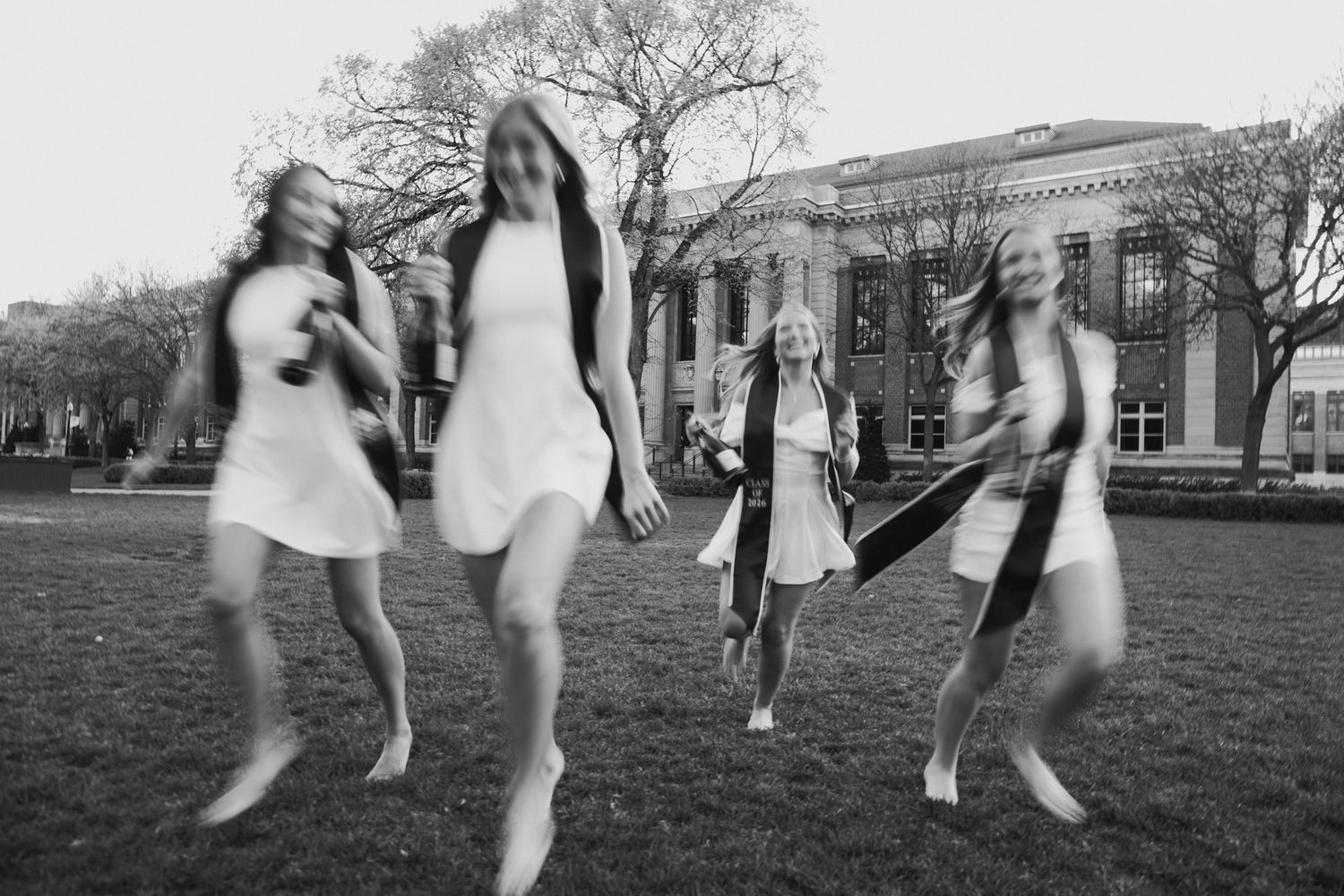 Black and white photo of four young women dancing joyfully on a grass lawn near a classic building.