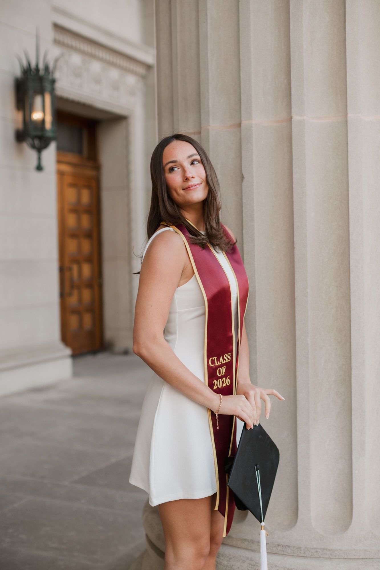 Brunette woman in white dress and maroon Class of 2026 graduation stole poses with cap by stone columns.
