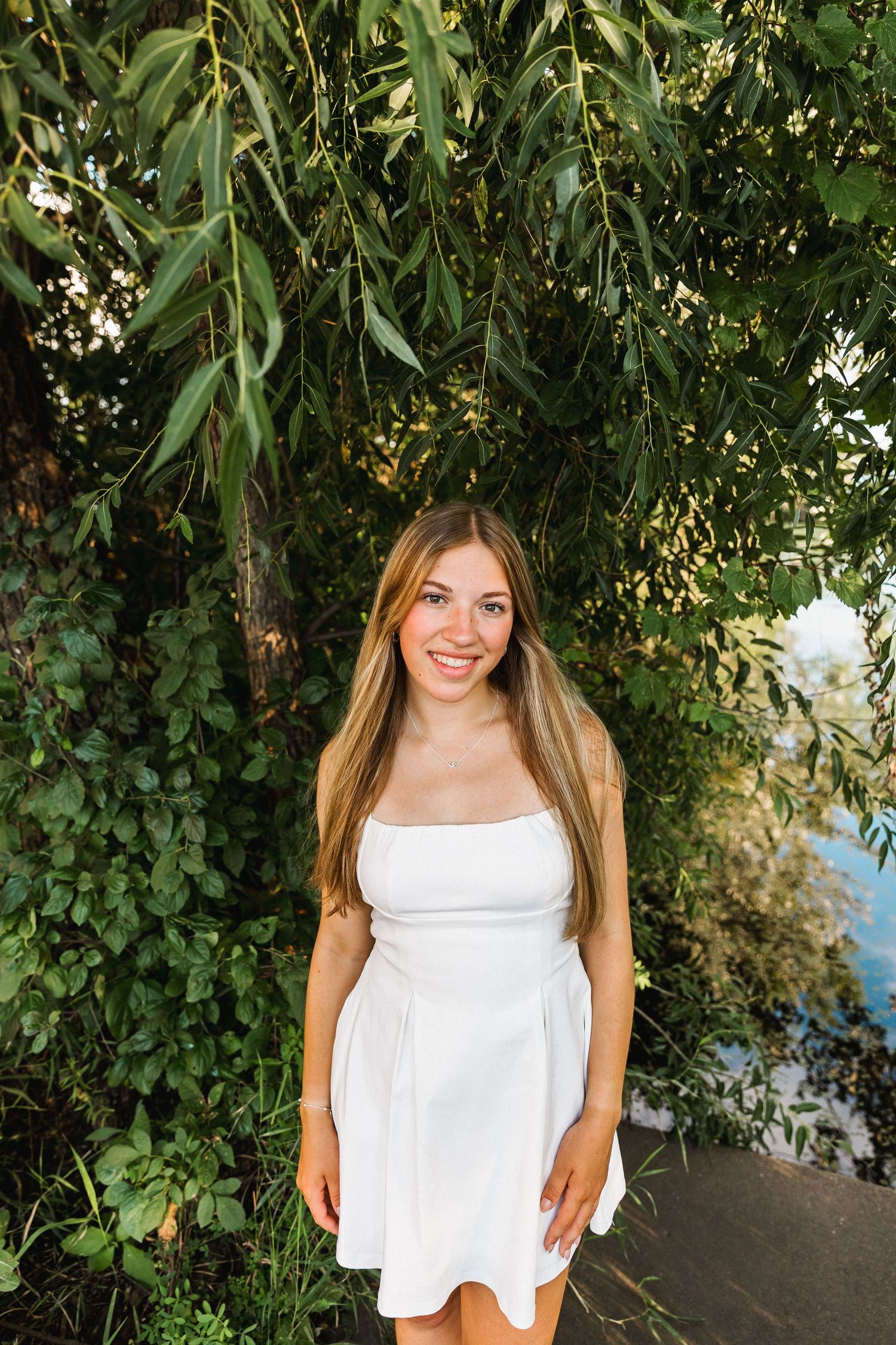 A person in a white sundress poses in front of lush green foliage on a sunny day.
