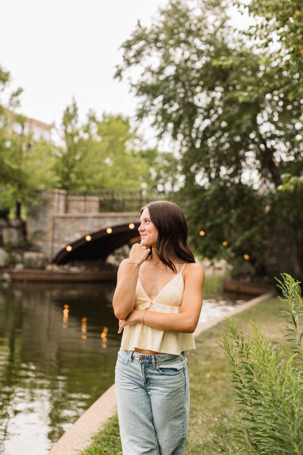 A woman in light yellow top and blue jeans poses near a scenic waterfront with trees and string lights at dusk.