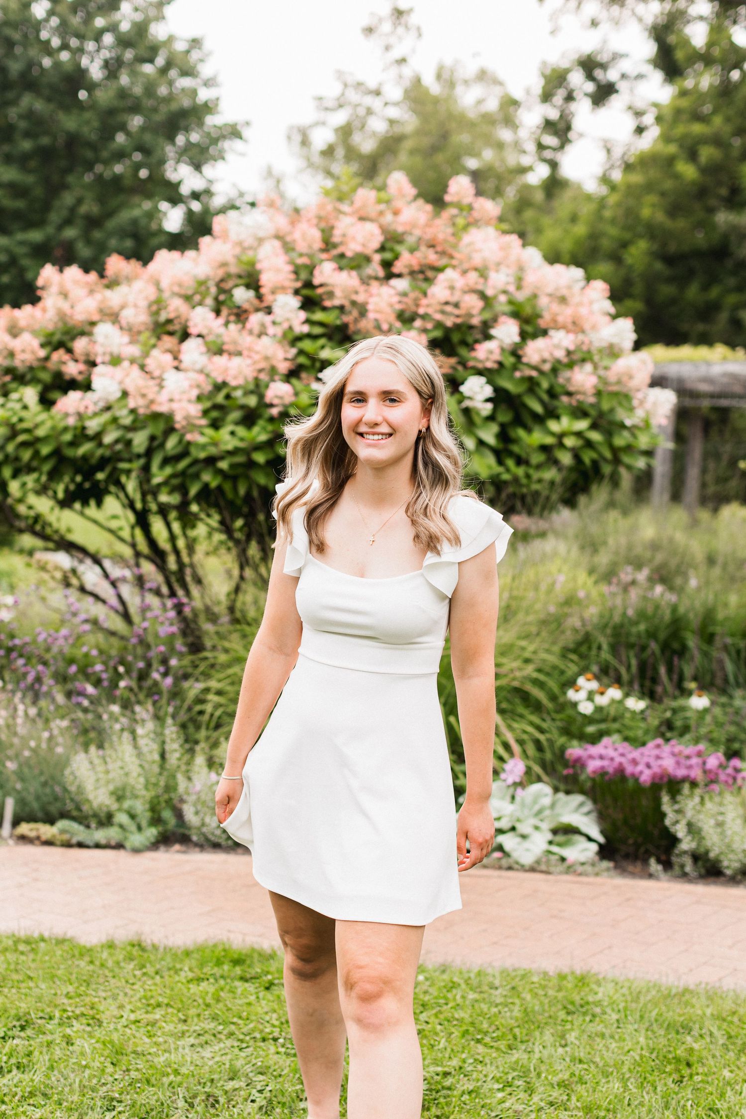 A white summer dress is worn outdoors in a garden with pink flowering bushes and greenery in the background.