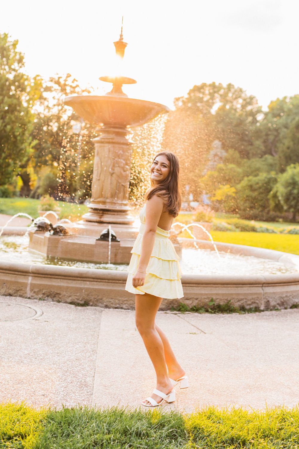 A child dances and spins in a yellow dress near a fountain during golden hour.