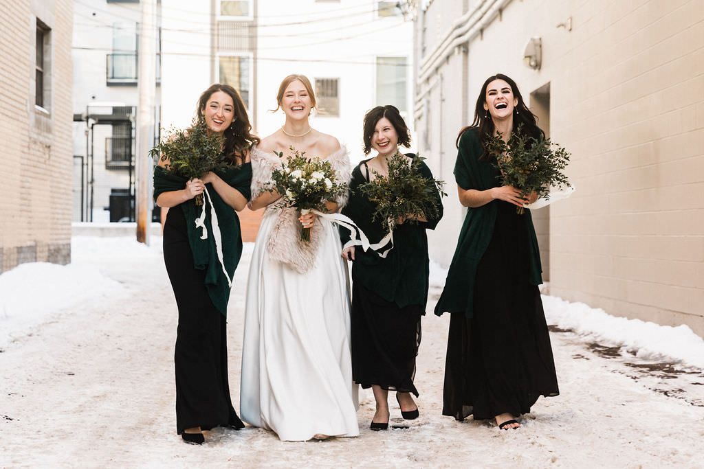 Wedding party stands together in winter snow wearing black dresses and white gown.