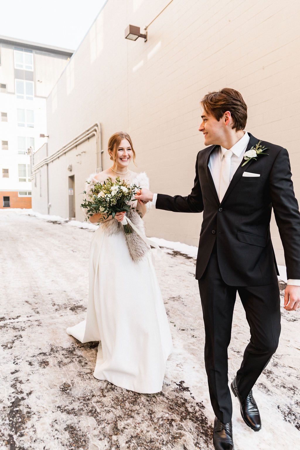 Newlyweds playfully walk through snow in formal wedding attire, creating candid winter moment.