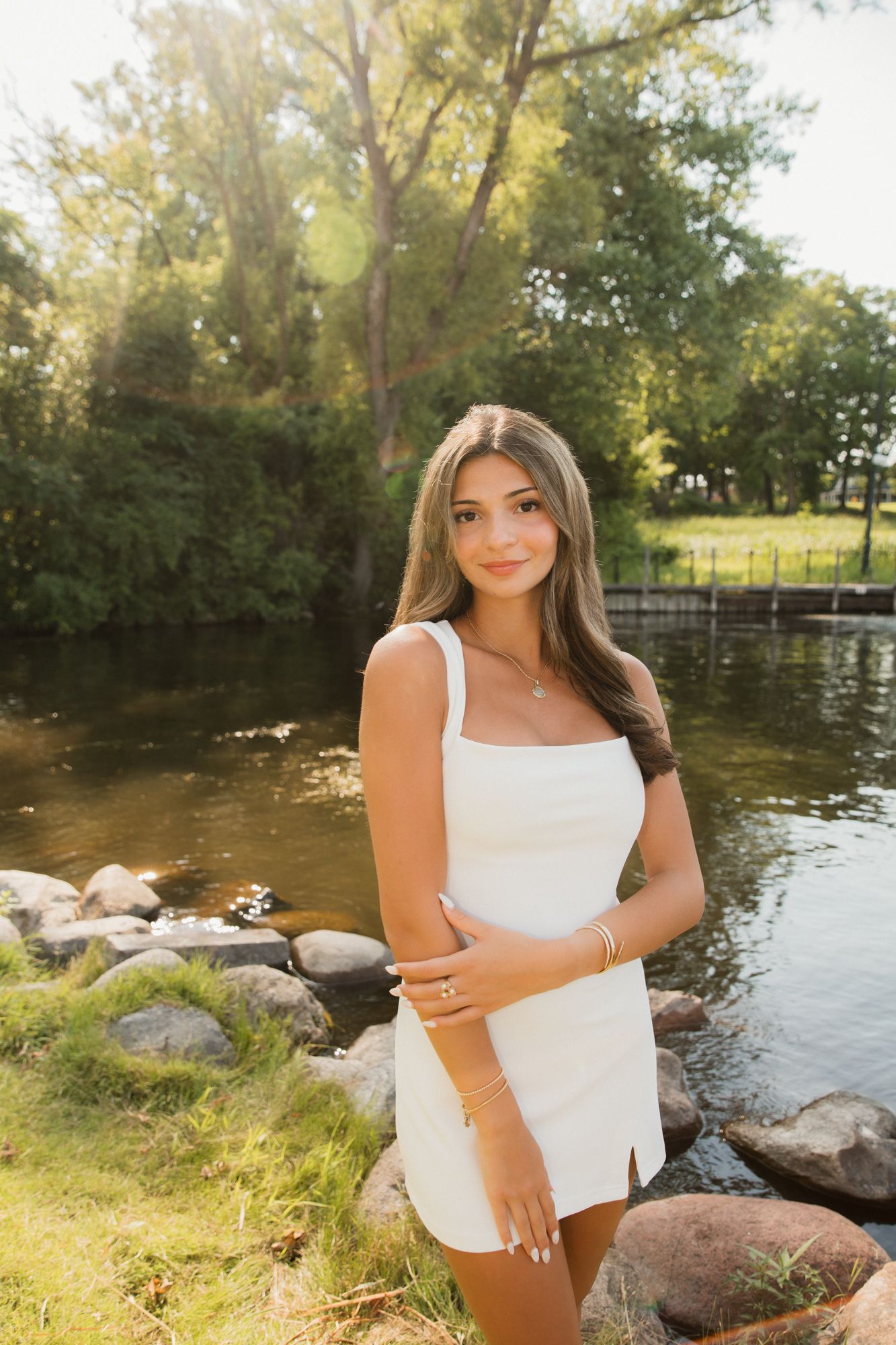 Young woman in white mini dress posing by a rocky riverside with lush green trees in golden sunlight.