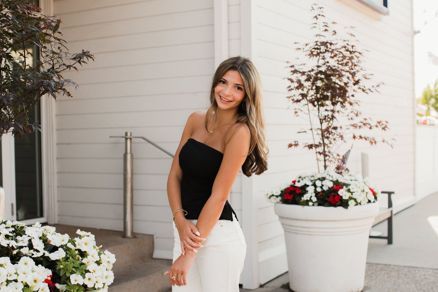 Young woman in black top and white pants posing outdoors near white planters with colorful flowers.