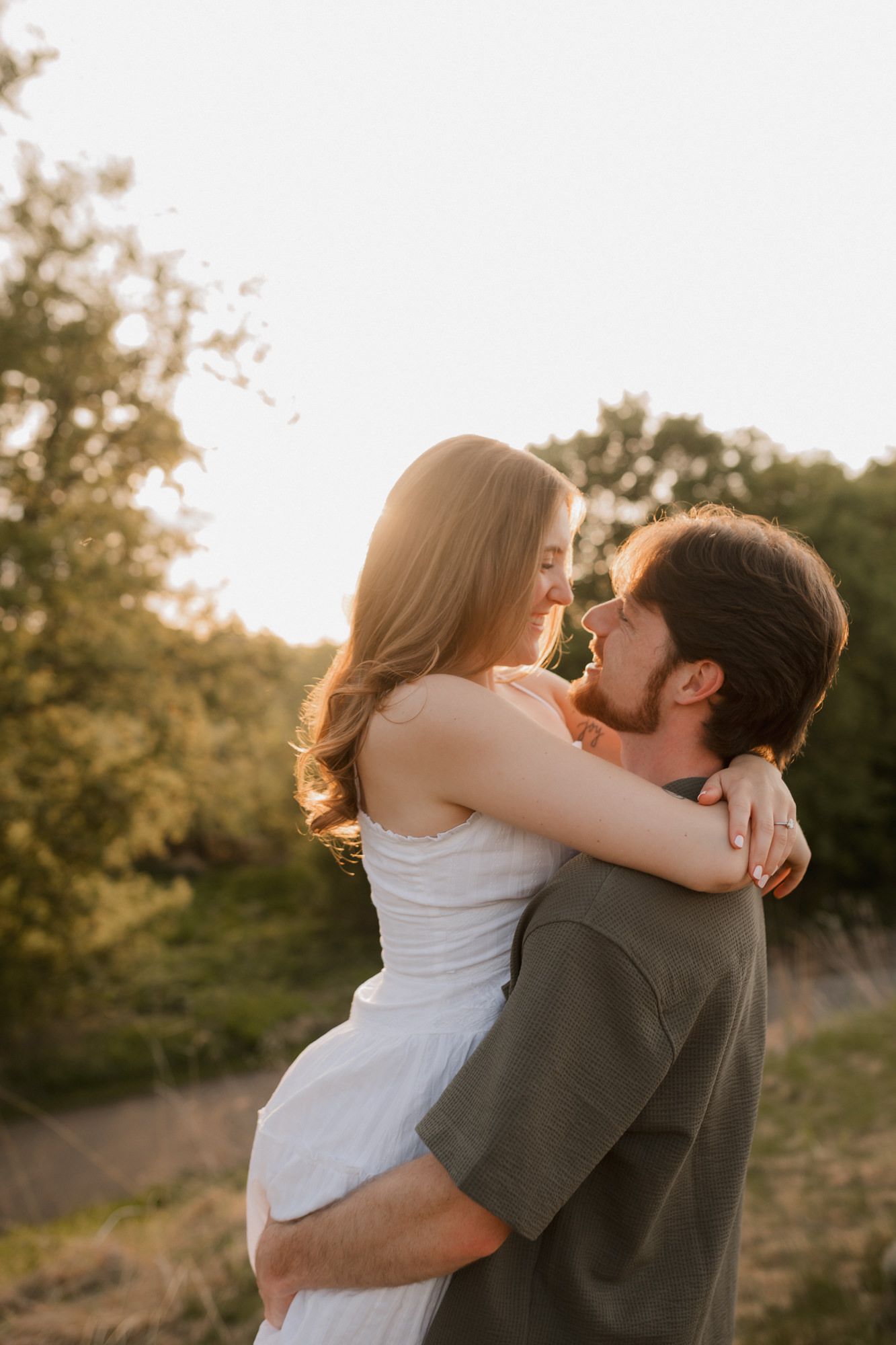 A couple embraces at golden hour, the man lifting the woman in a white dress outdoors surrounded by lush green trees.