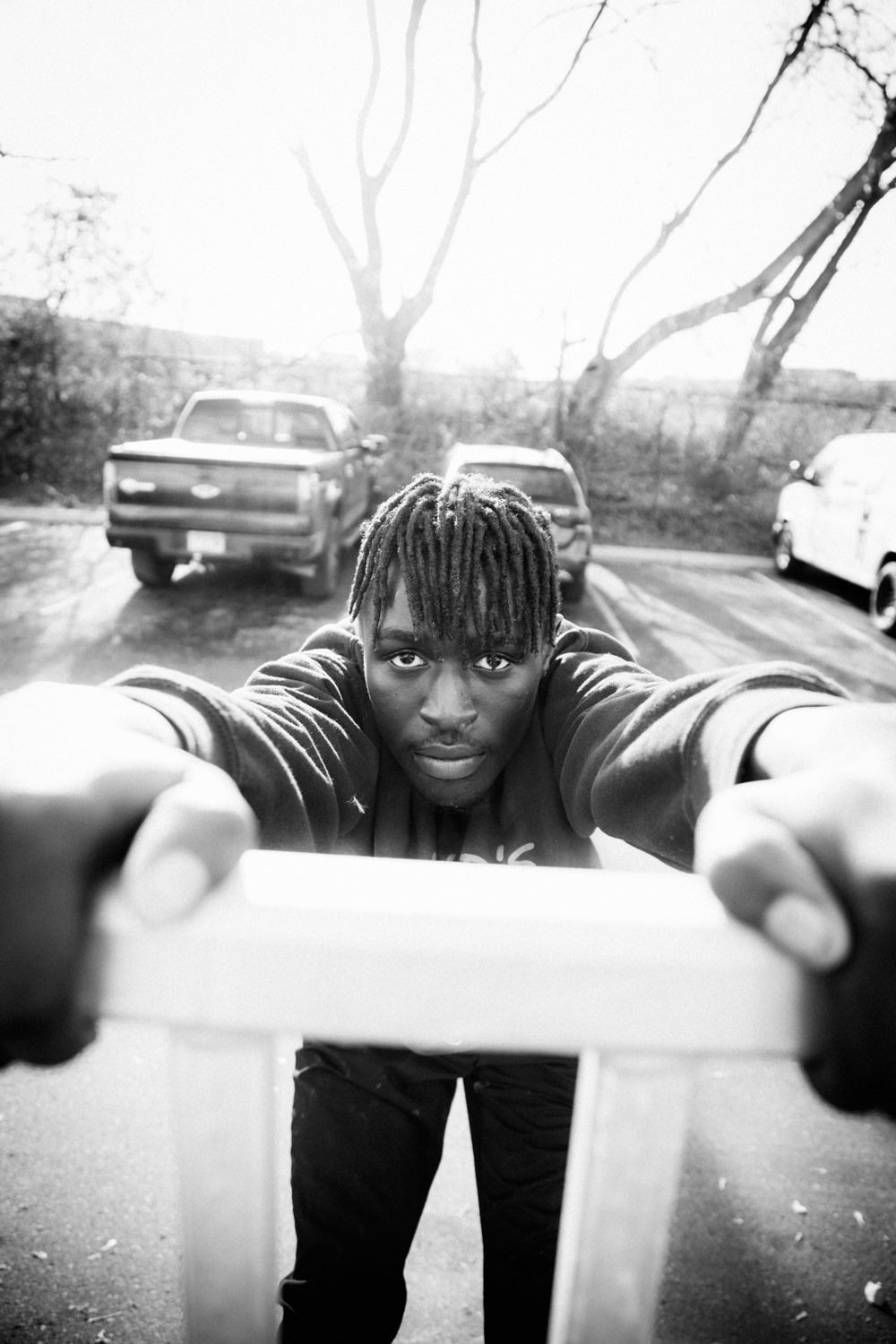 Black and white photo of a young man with dreadlocks leaning toward camera in a parking lot.