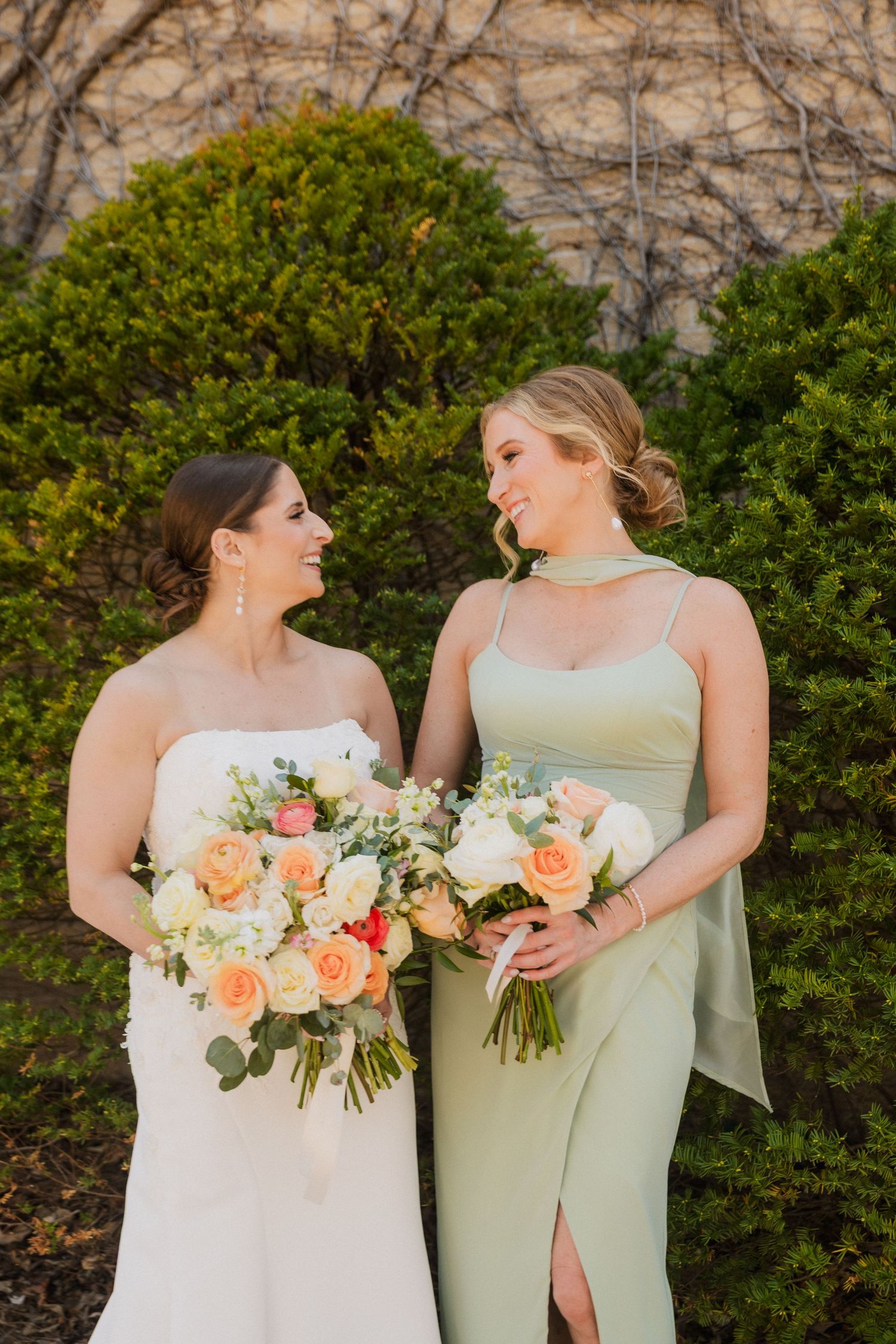 Bride in white strapless gown and bridesmaid in sage green dress holding peach rose bouquets, smiling outdoors.