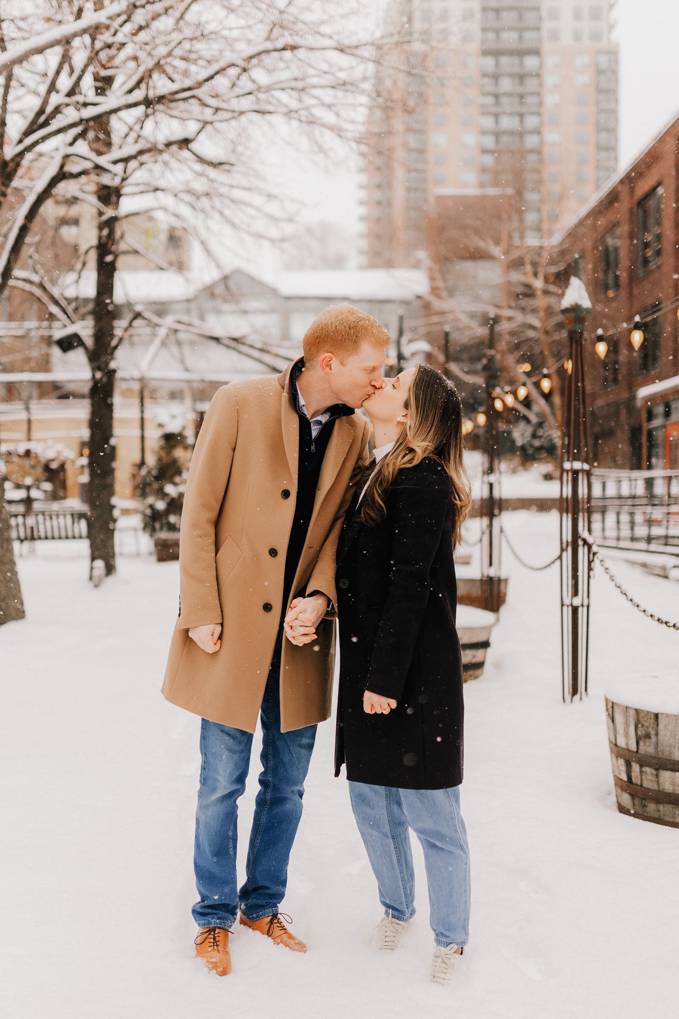 Couple kissing in snowy urban setting, wearing elegant winter coats while holding hands with city lights in background.