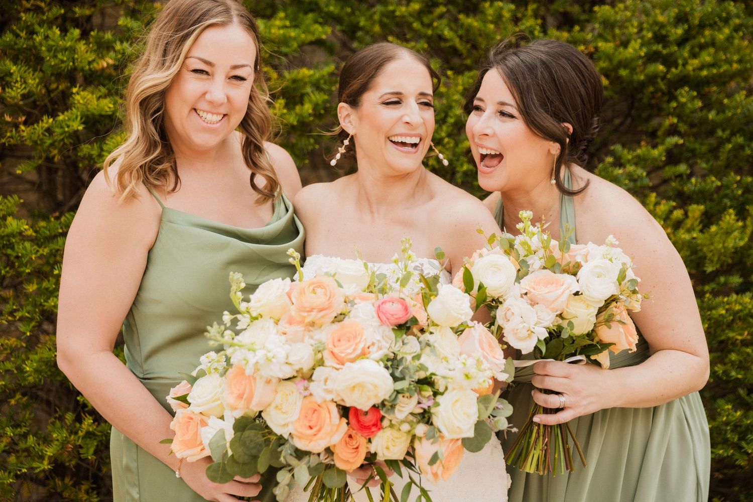 Laughing bride in white gown flanked by two bridesmaids in sage green dresses holding peach and white bouquets.