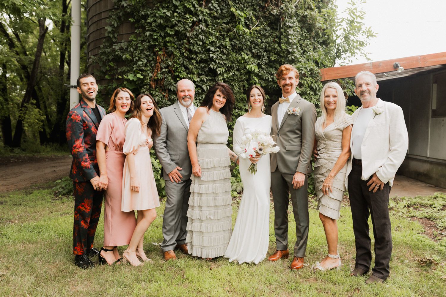 A wedding party in blush pink and gray attire poses together for outdoor group photos in a garden setting.