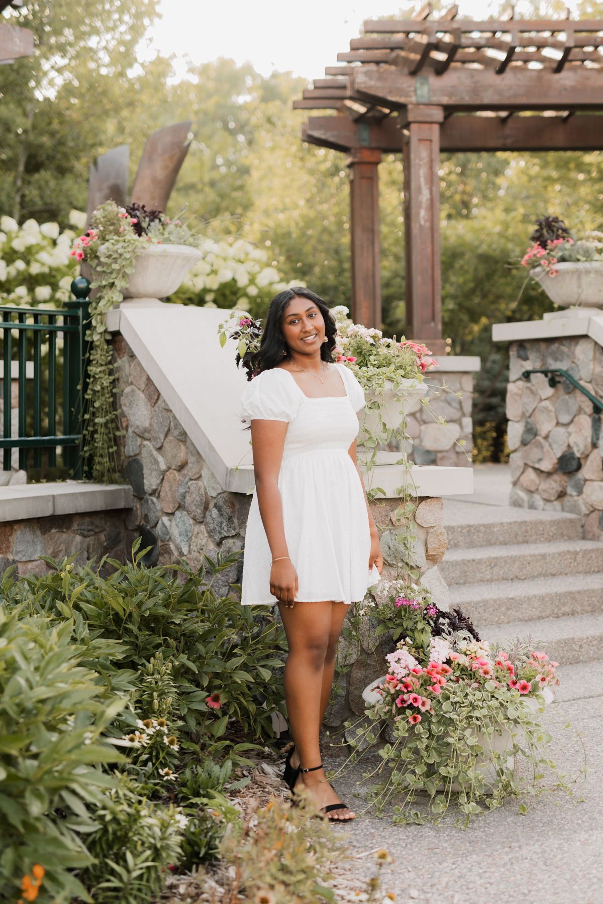Young woman in white mini dress posing in a garden with stone steps and colorful flowers.