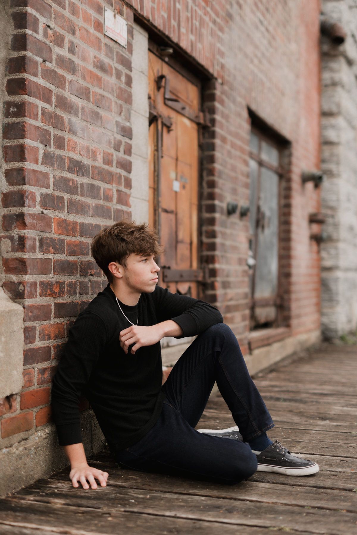 Young man in black outfit sitting against brick wall on wooden deck in urban alley setting.