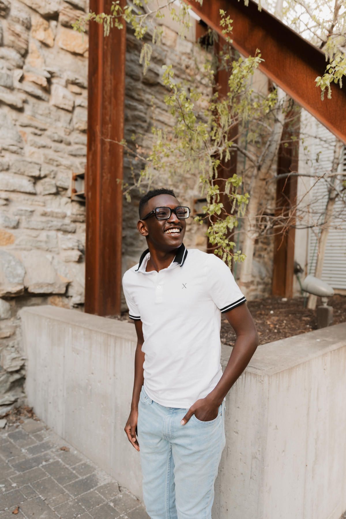 Young Black man in white polo shirt and glasses smiling outdoors near stone wall and rustic beams.