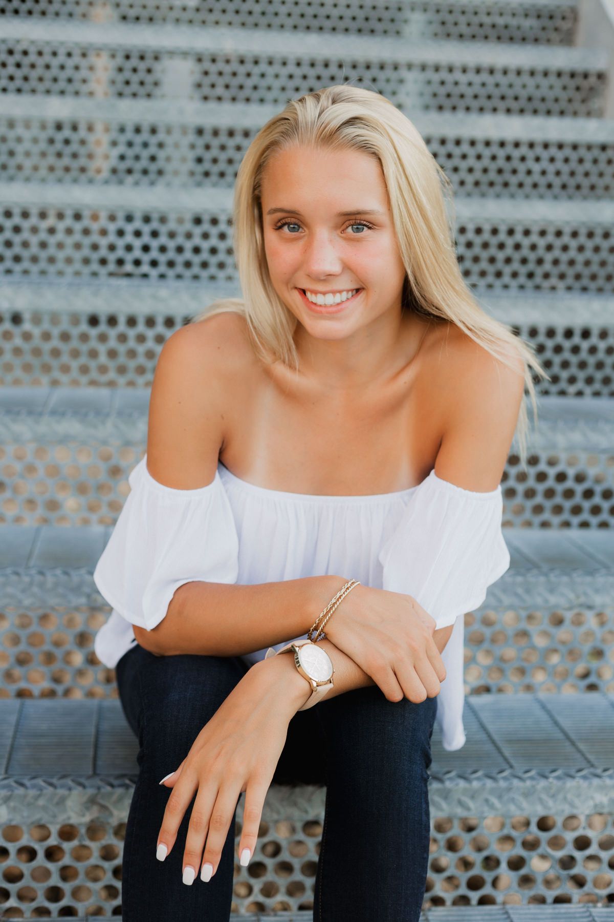 Smiling blonde girl in white off-shoulder top sitting on metal steps, wearing a watch and bracelet.