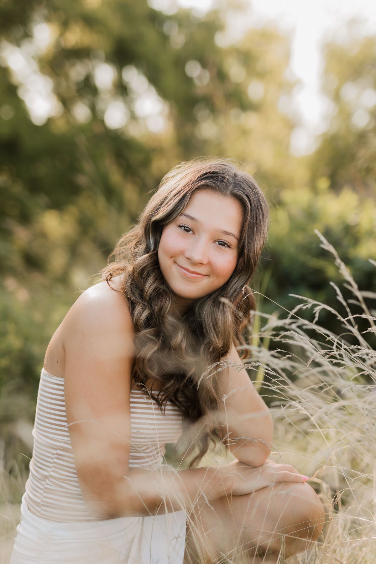 Young woman with wavy brown hair smiling in a golden field of tall grass during sunset.