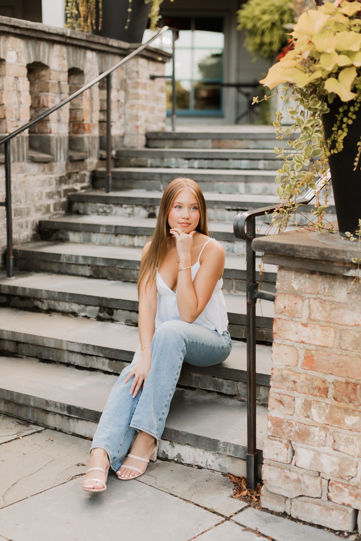 Young woman with long hair sits on stone steps in white tank top and jeans, posing thoughtfully outdoors.