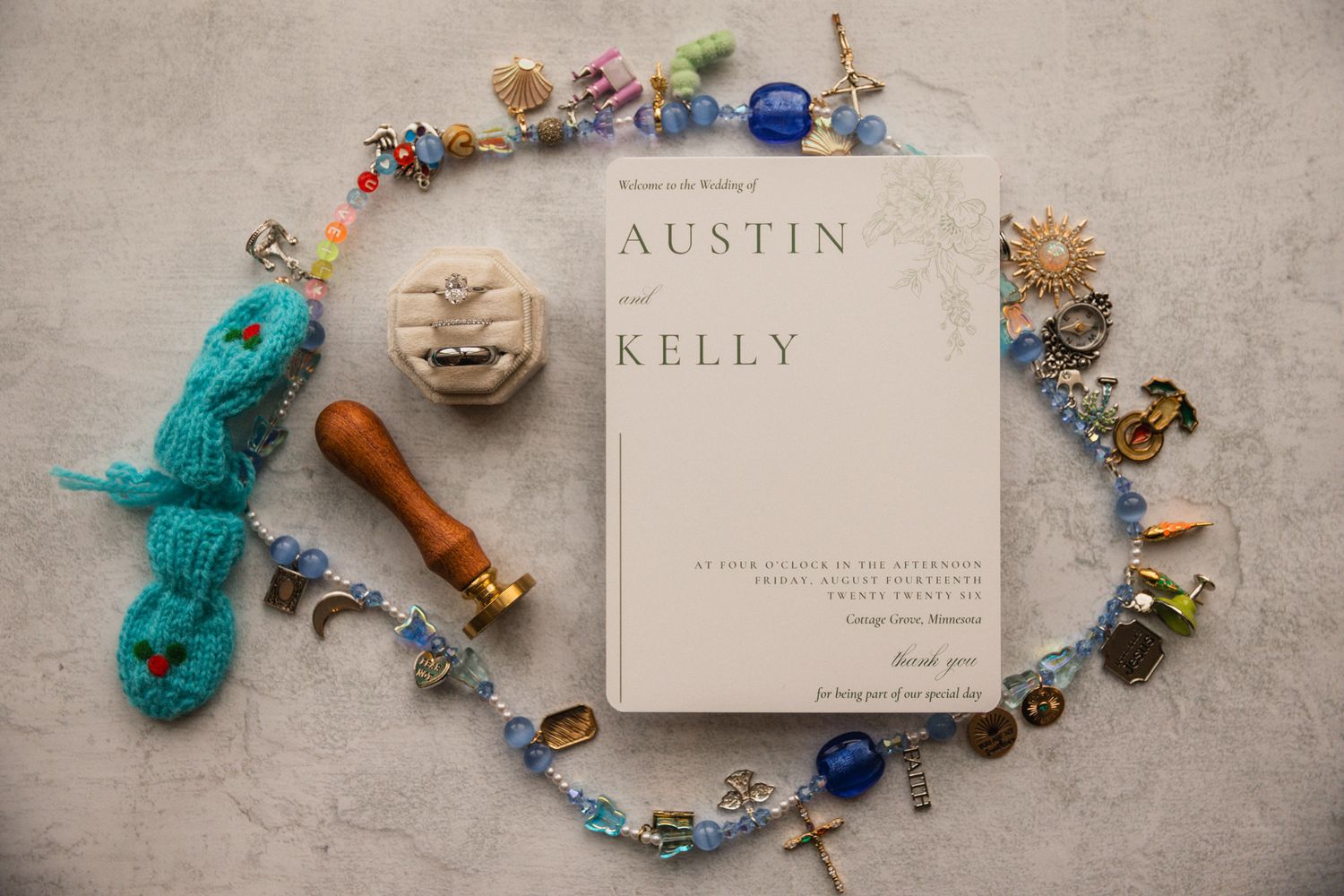 Austin and Kelly wedding invitation surrounded by colorful beaded jewelry, charms, and a teal knitted accessory on marble.