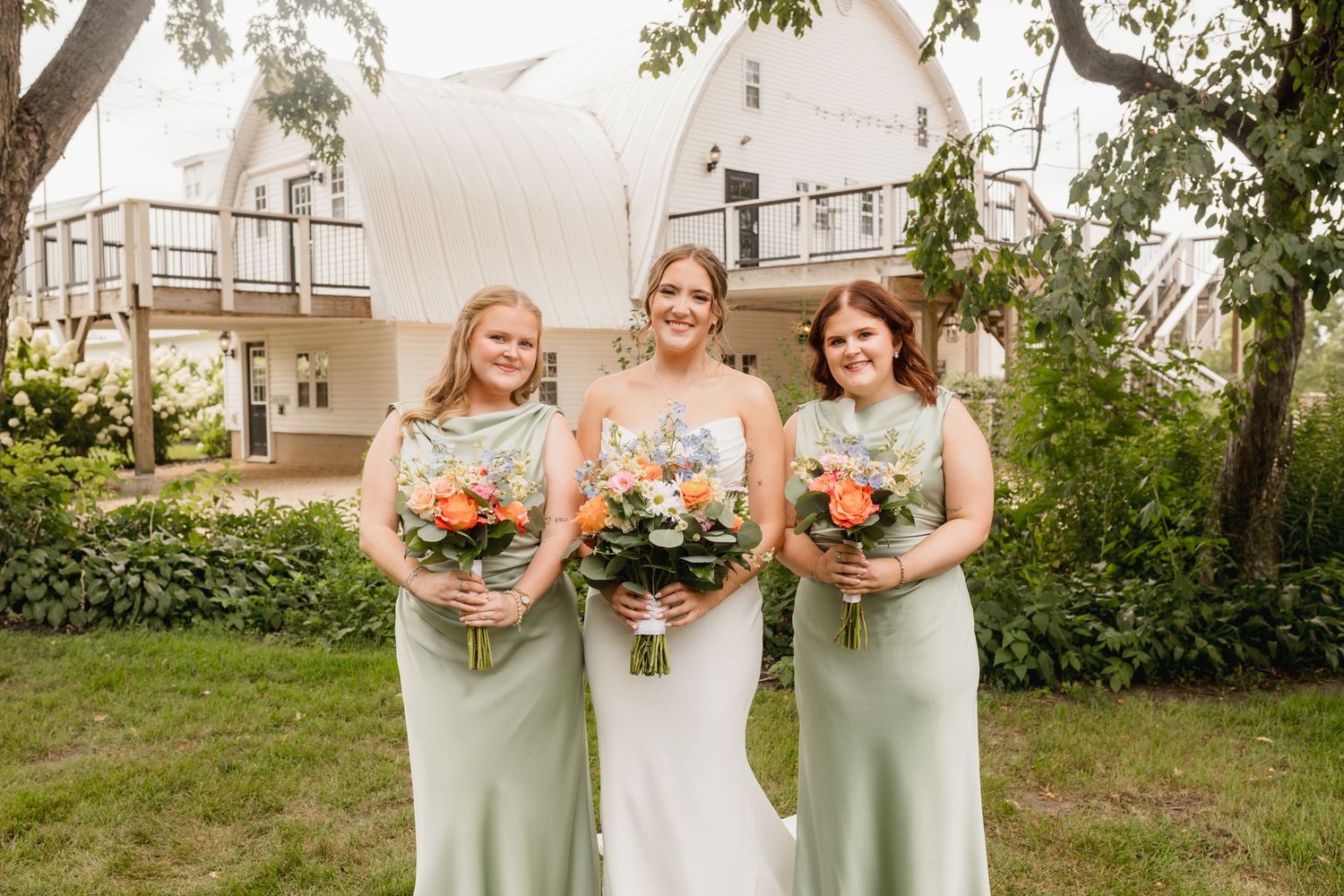 Bride in white gown flanked by two bridesmaids in sage green dresses holding colorful bouquets outdoors.