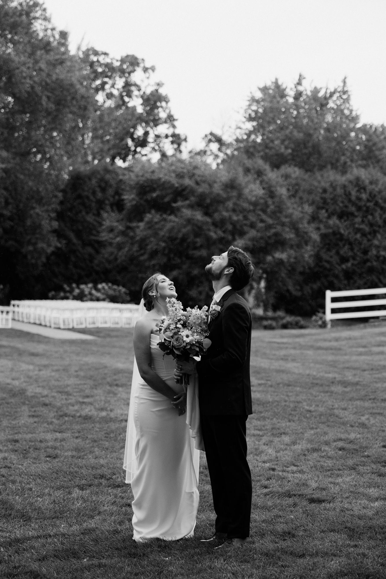 Black and white photo of a bride and groom standing on a lawn, both looking up, surrounded by lush trees.