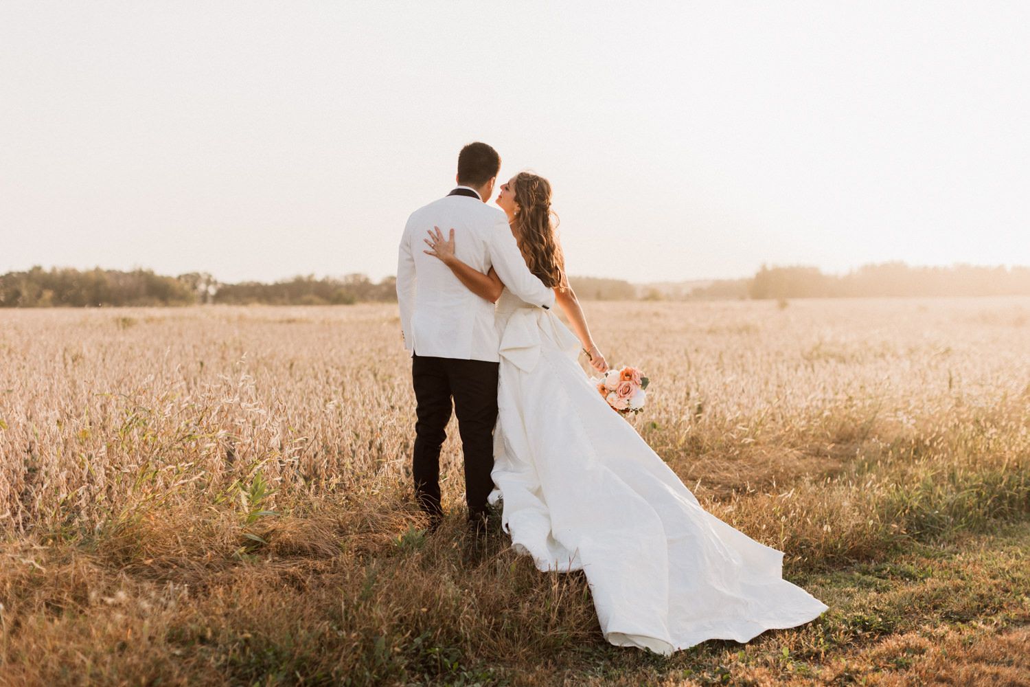 A couple in wedding attire embraces in a golden sunlit field during sunset.