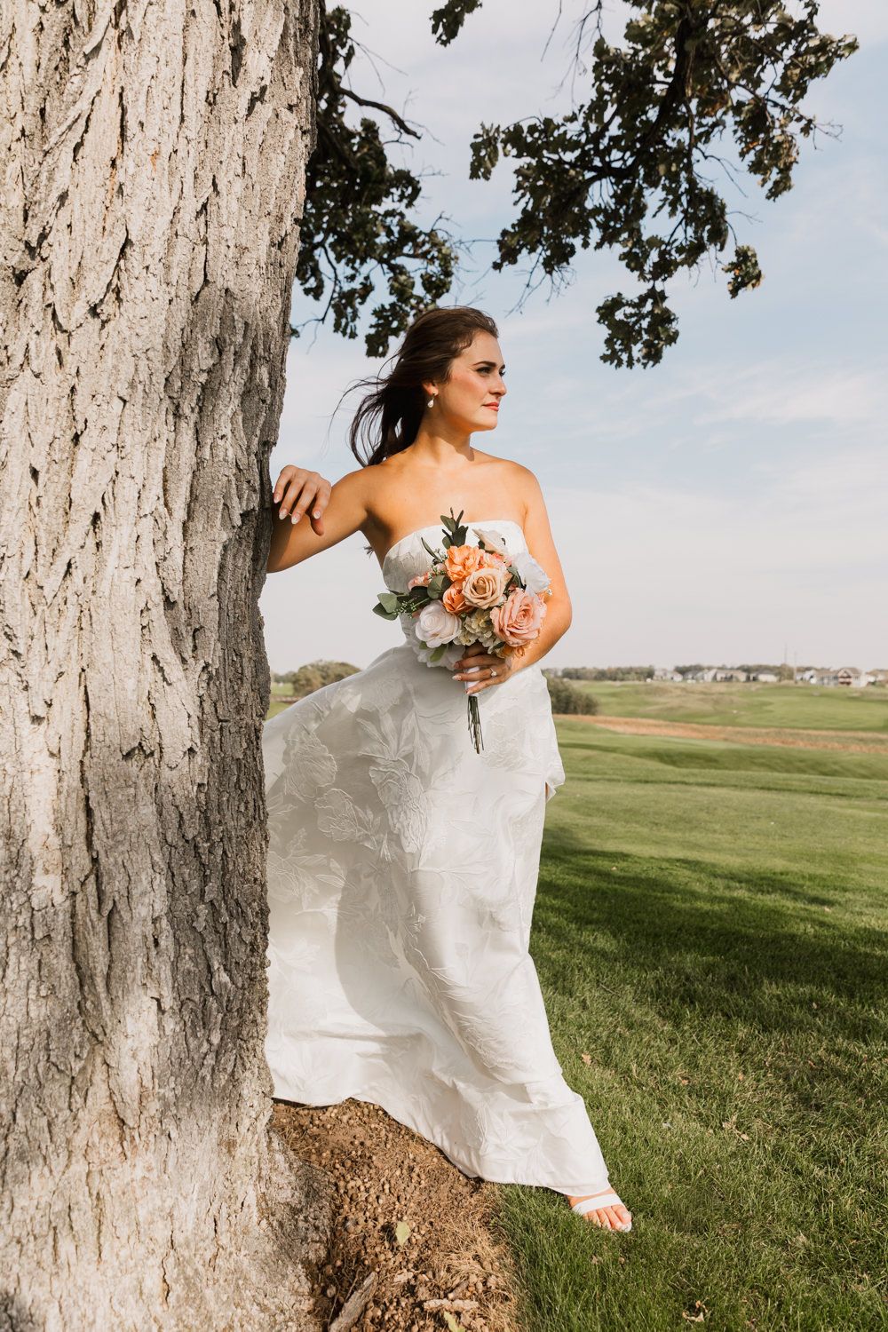 A bride in a white wedding dress poses gracefully near a large tree trunk in a grassy field on a sunny day.