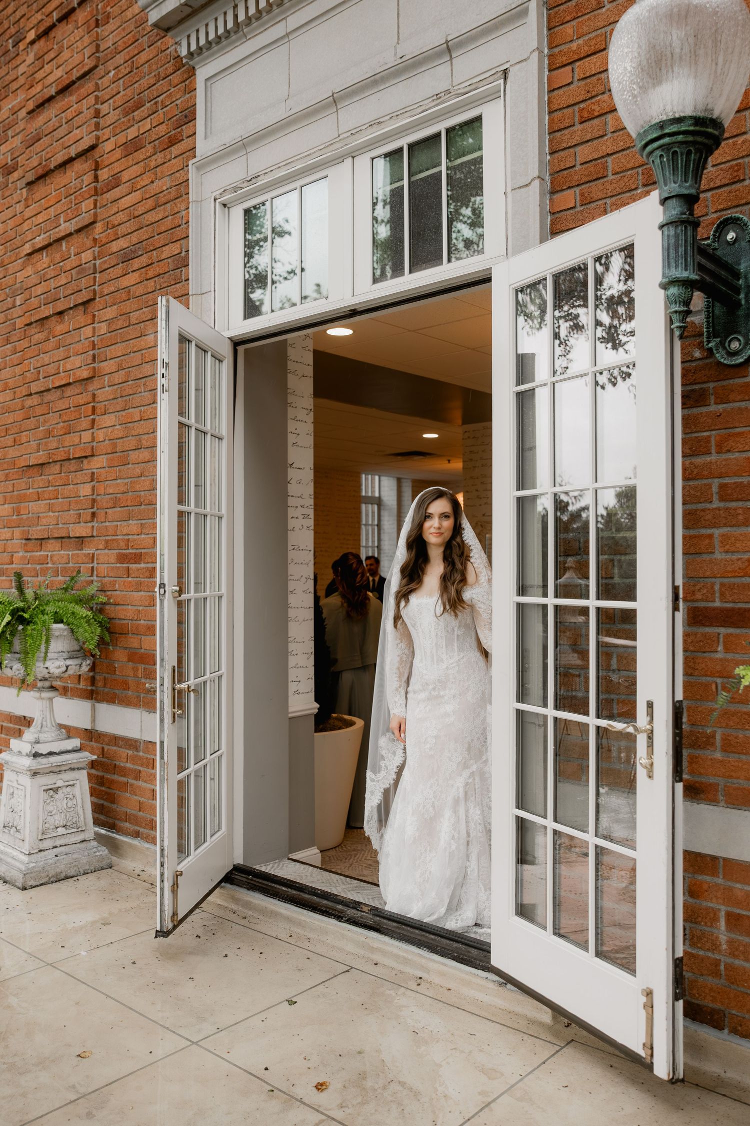 Bride in lace wedding gown and veil standing in open French doors of brick building.