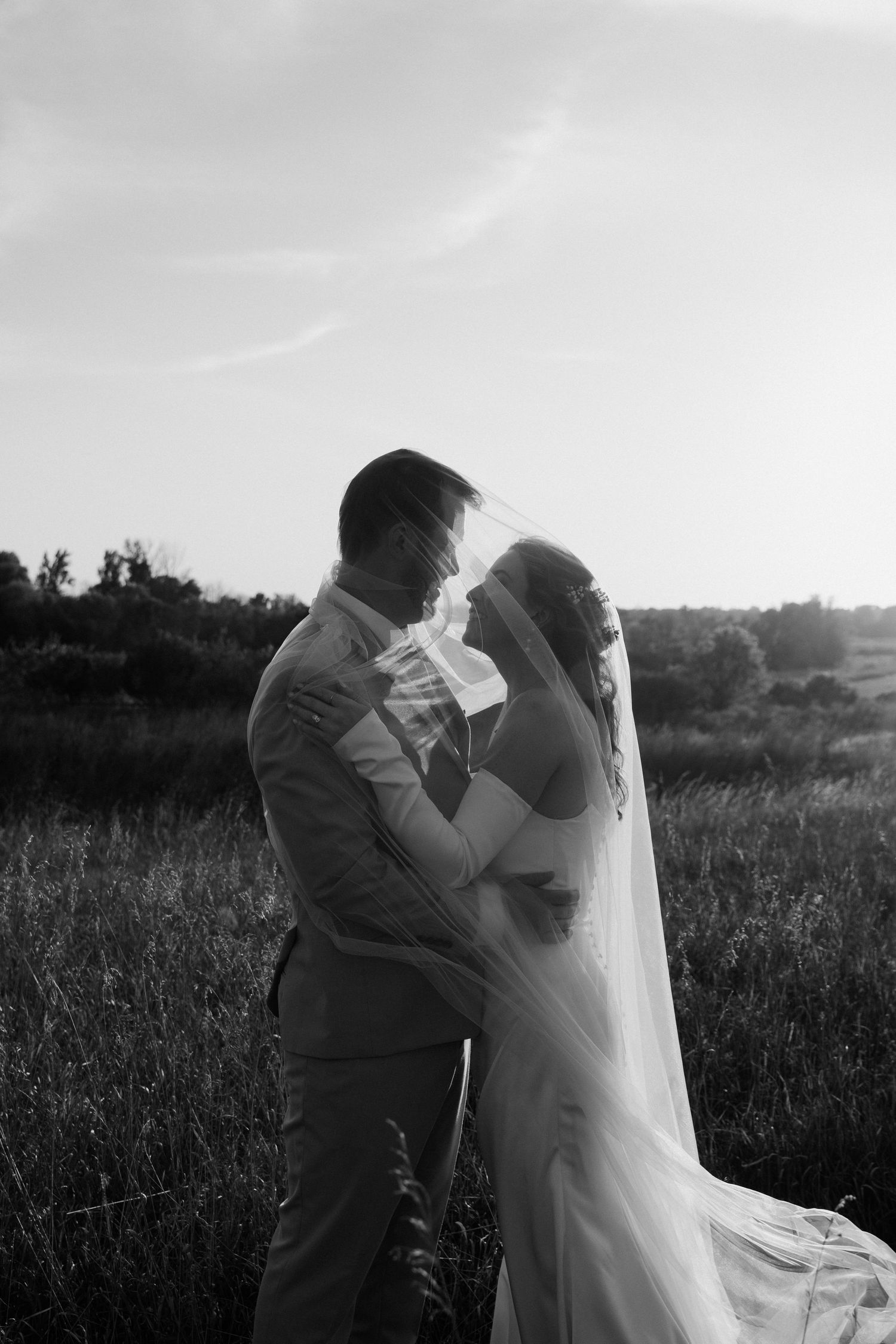 Black and white photo of bride and groom sharing romantic moment under veil in field at sunset.