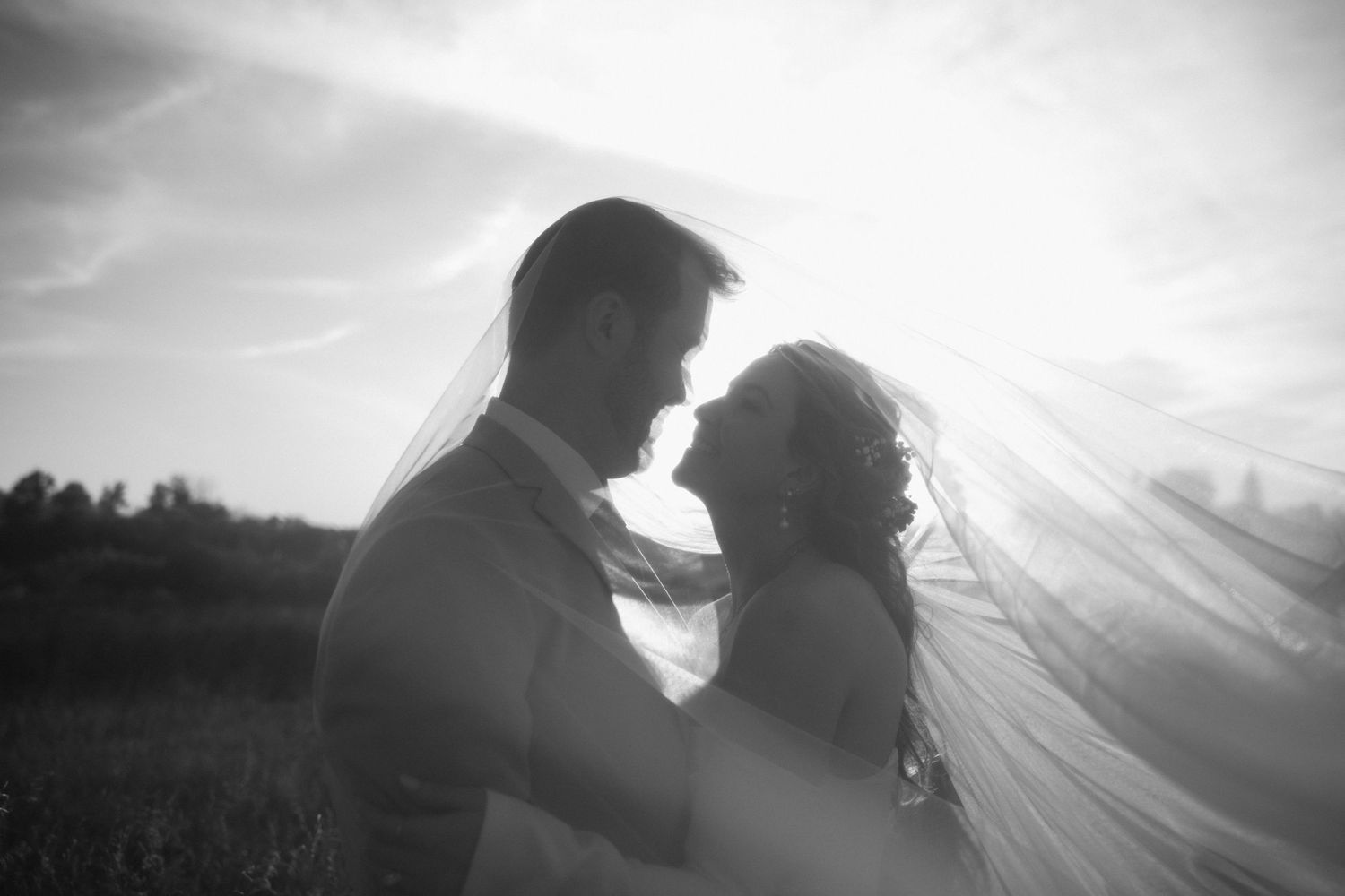 Black and white image of a bride and groom nearly kissing beneath a flowing veil against a bright sky.