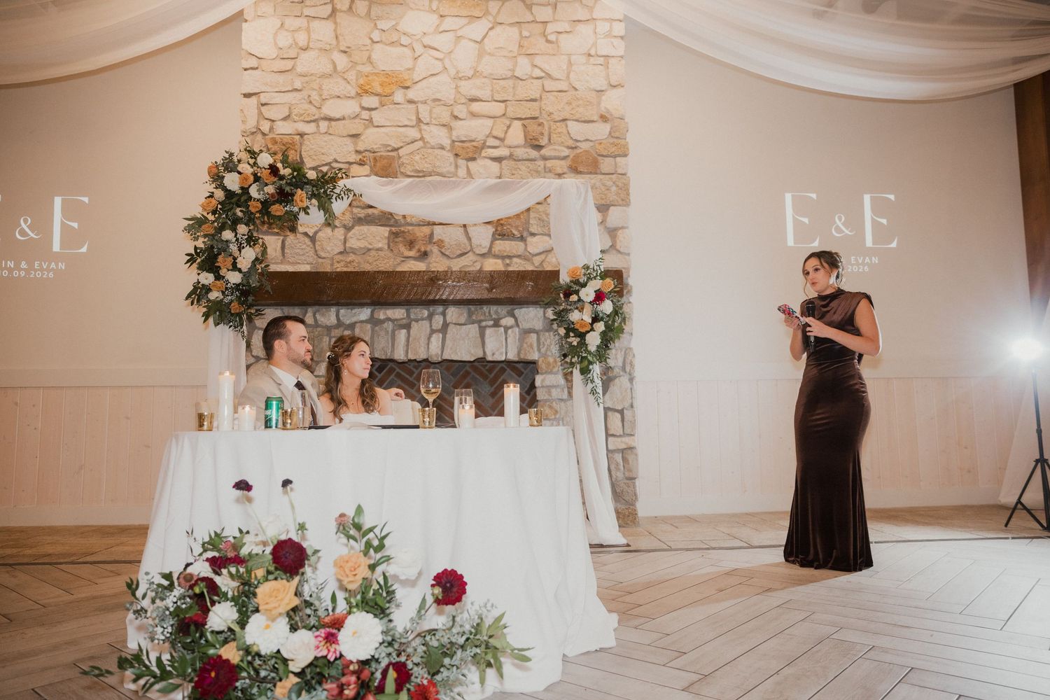 Bridesmaid in a burgundy gown gives a wedding toast beside a sweetheart table with floral arrangements and a stone fireplace.